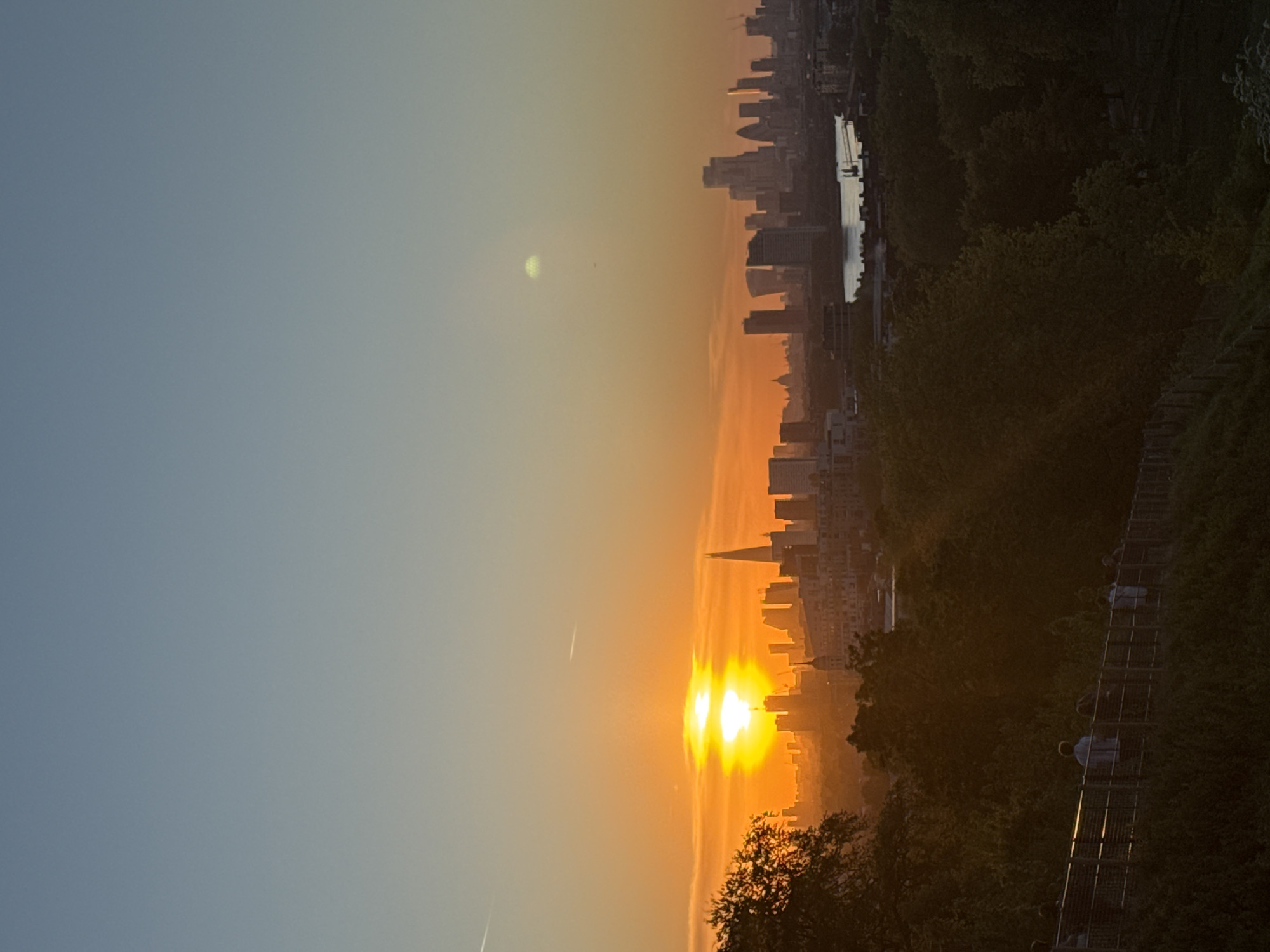 The image, when oriented correctly, captures a panoramic cityscape at either sunrise or sunset, dominated by warm, golden light. The sky transitions from a soft blue at the top to vibrant orange and yellow hues near the horizon, where the sun is a prominent and intense feature, appearing somewhat diffused and creating lens flares. Faint contrails are visible in the upper sky.
A dense skyline of modern buildings stretches across the midground. A distinct, pointed skyscraper, strongly resembling The Shard, is visible near the brightest part of the sun's glow, amidst other high-rise structures. Behind sections of the city, a body of water, likely the River Thames, reflects the bright sky.
In the foreground and extending to the right, a dark, heavily wooded area of trees and foliage forms a natural boundary, suggesting a park or elevated green space. On the far right, a long, sloping structure, appearing to be a staircase or ramp with railings, descends into this wooded area. Several human figures are visible on this structure, appearing to be walking. The trees are largely silhouetted against the radiant horizon. The scene provides an expansive and serene view of London during a golden hour. No visible text is present.