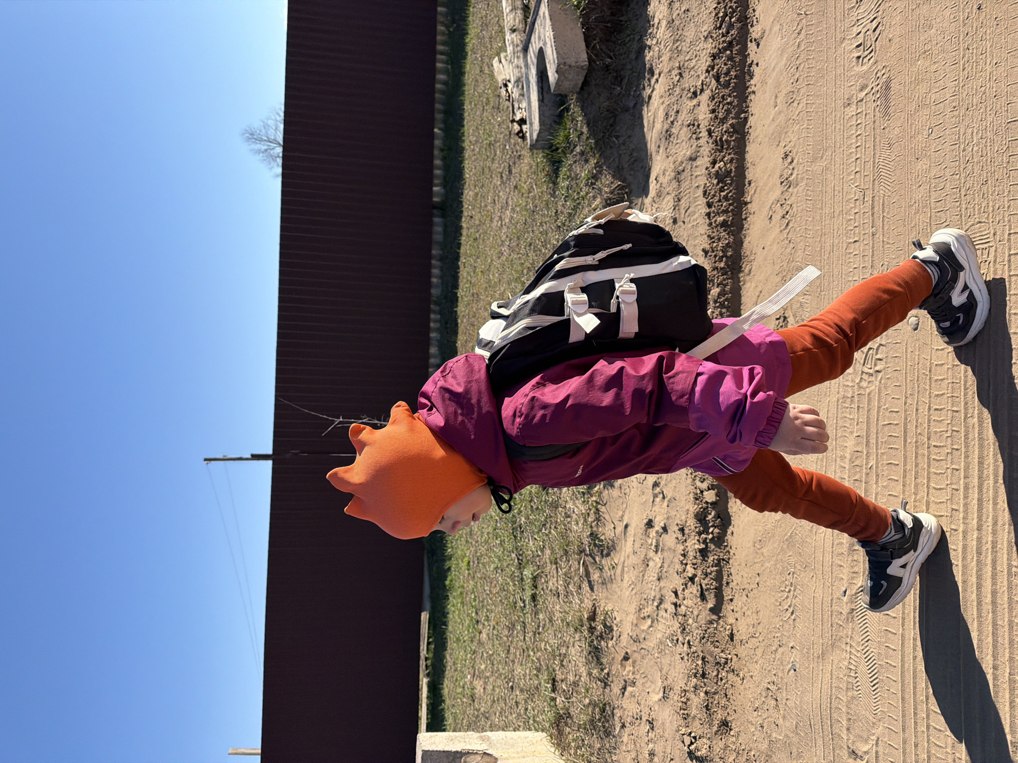 A child wearing a red-orange hat, reddish-purple jacket, and rust-colored pants is walking on a dirt path. The child has a black and white backpack on. Tire tracks are visible on the path. A metal fence is on the left side of the frame, and a clear blue sky is above. The picture was taken in Chaikovsky, Russia.
