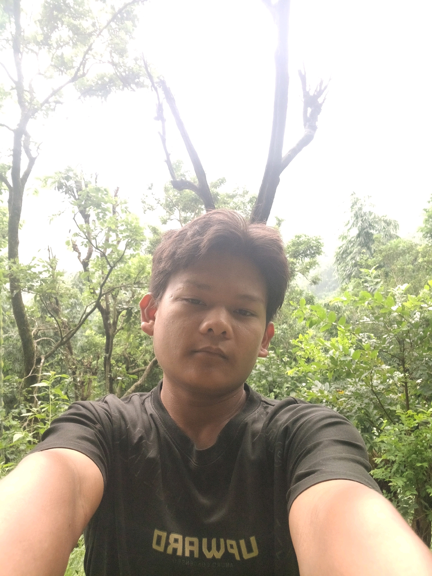 A young person with short, dark hair takes a selfie in a forest. They are wearing a dark t-shirt with a faded logo that appears to read "FORWARD". The background is filled with lush green trees and foliage, suggesting a natural, outdoor environment. The lighting is bright, indicating it might be daytime. The person's face is centrally located in the frame, and they are looking directly at the camera. The image appears to have been taken in the city of Triyuga, Nepal, a location known for its natural beauty and forested areas.