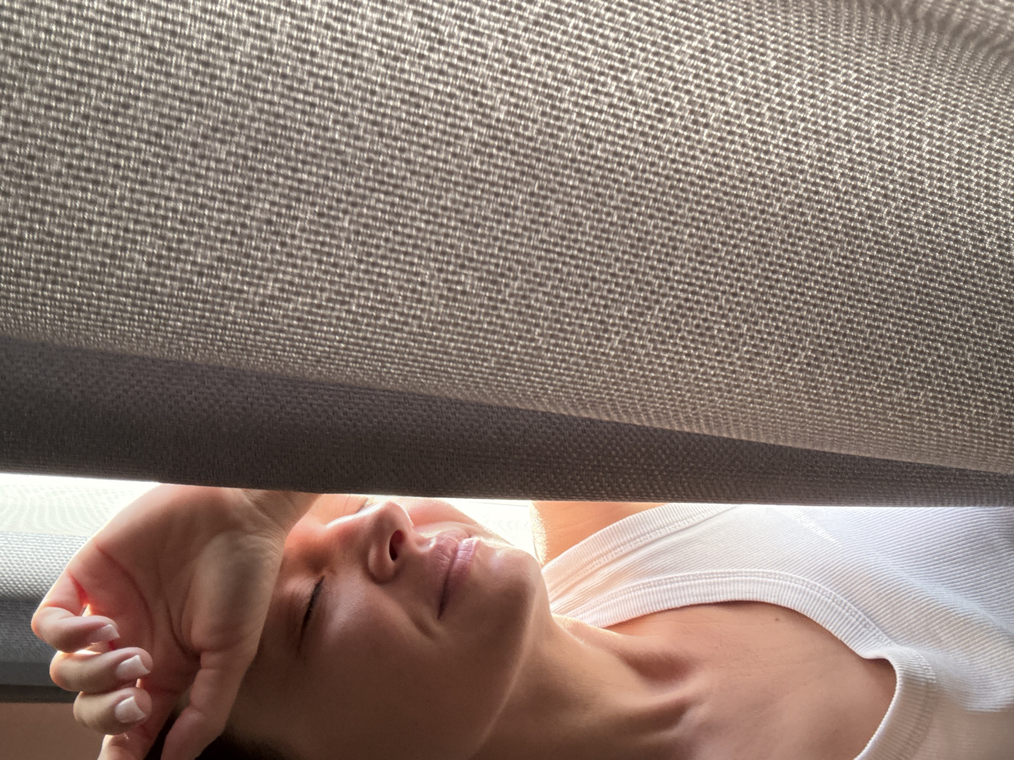 A woman lies on her back, her right hand resting on her forehead. She is wearing a white tank top. Above her is a textured, gray fabric, possibly a car sunshade. The location is indiscernible; no details reveal that this image was taken in the city of Chaikovsky, Russia.