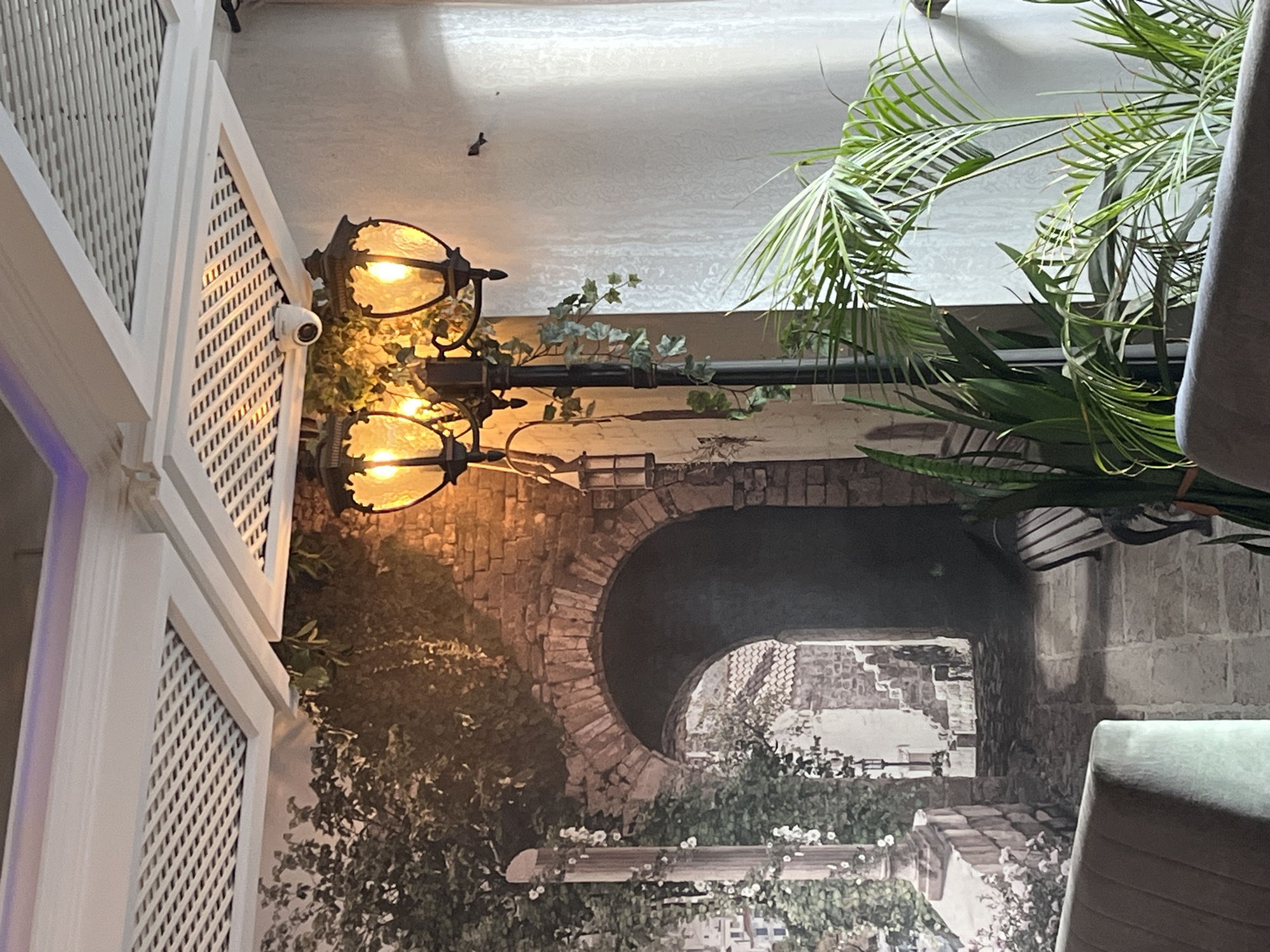 The image depicts an indoor scene with elements suggesting a restaurant or decorative space. A white latticework structure is on the left, with a security camera mounted. A vintage-style lamppost with two lit lamps is attached to the structure. The background features a mural of a brick archway leading to a European-style building with foliage. A potted palm is partially visible on the right. The location cannot be verified as being in Burgas, Bulgaria from the image provided.