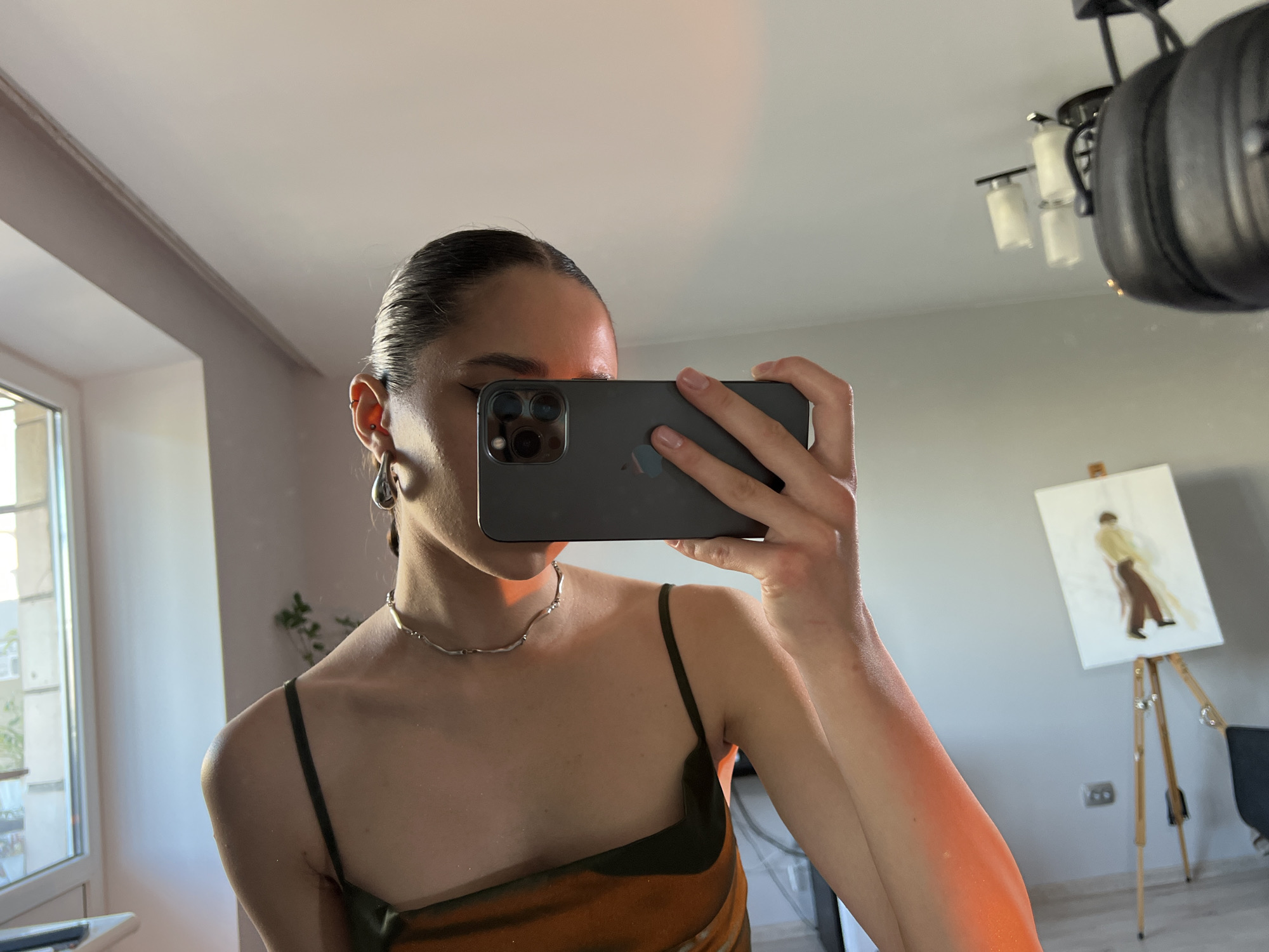 A fair-skinned individual is holding a smartphone in front of their face, obscuring the central portion of their face. Only the upper forehead, eye, and lower portion of the face are visible. The person has their hair pulled back from their face. They are wearing teardrop earrings and a silver necklace. They wear a spaghetti-strap top or dress.
The scene appears to be indoors, with a window partially visible on the left. In the background to the right, there's an easel with a painting of a person standing. A ceiling fan/light fixture is visible in the upper right corner. It is not possible to determine the location in the city of Ternopil, Ukraine from this image.