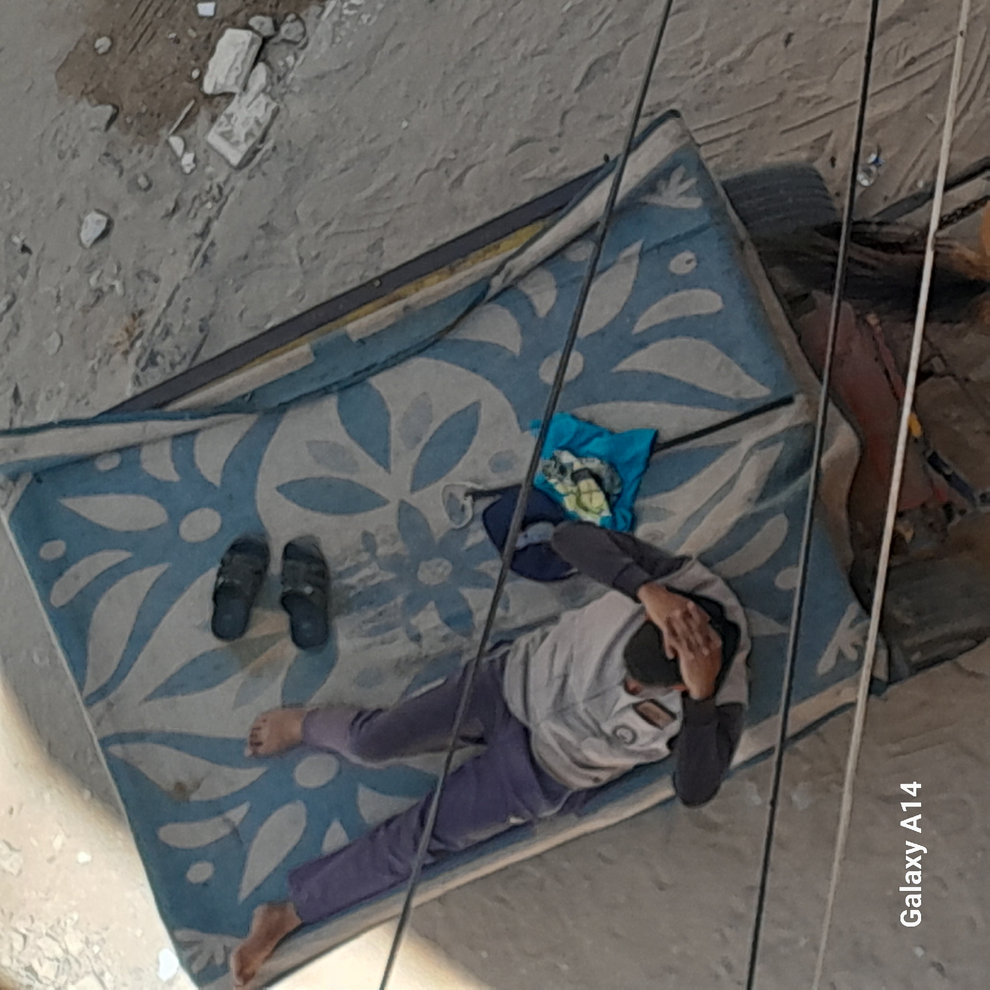 An aerial shot shows a person lying on a mattress on top of a cart in Khan Yunis, Palestinian Territories. The mattress has a blue and white floral pattern and is slightly soiled. The person is wearing dark purple pants and a light gray sweatshirt, with a black beanie covering their head. They are lying on their back, with their legs extended and their head resting on their hands. Two dark-colored sandals are placed on the mattress near the person's feet. A folded blue cloth and what appears to be some debris are situated near the person's head. The cart has a metal frame and appears to be in a dusty outdoor environment, with debris scattered on the ground. Black wires are visible in the foreground, obscuring parts of the scene. The bottom left corner of the image displays the text "Galaxy A14".