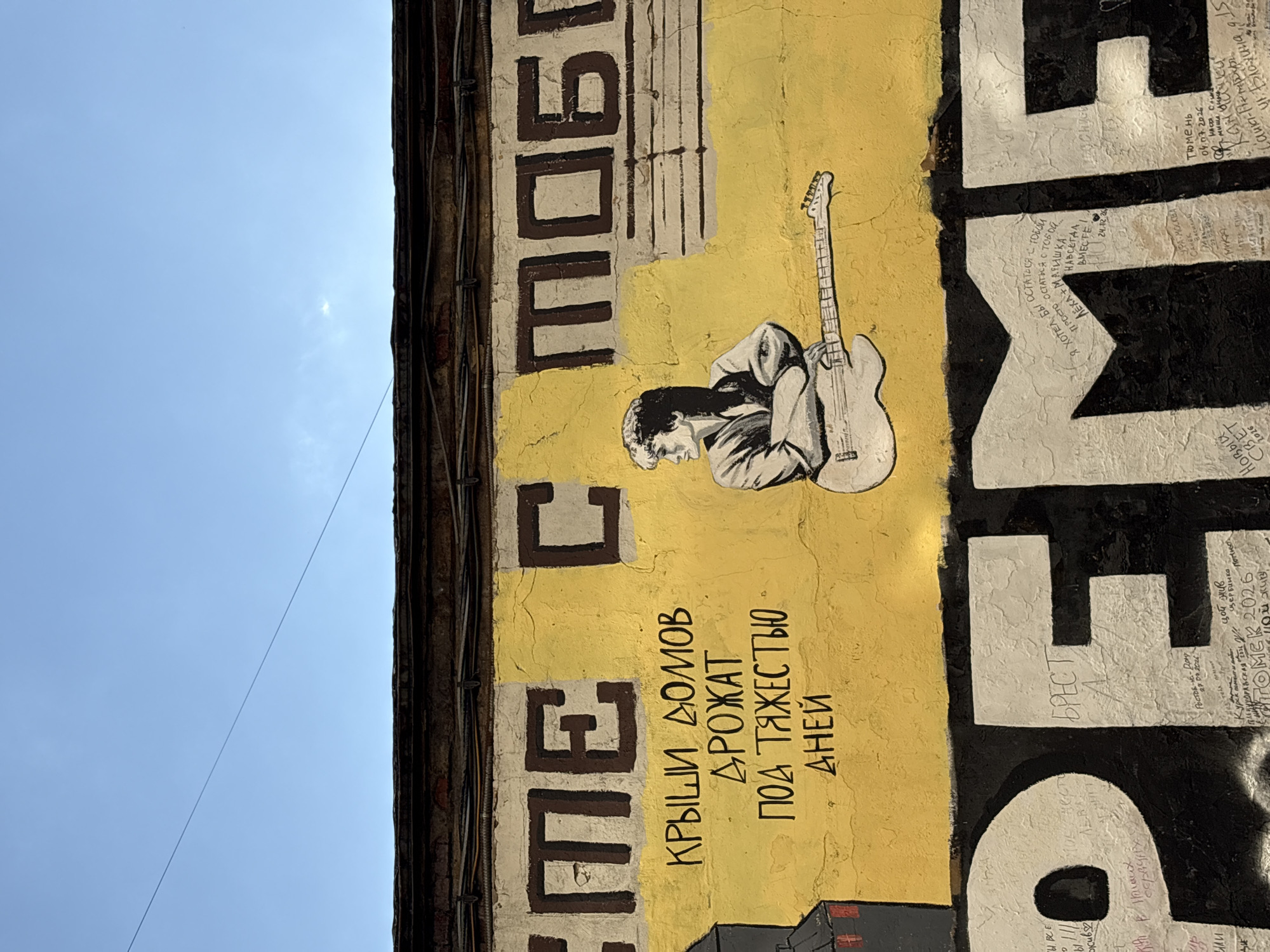 The image shows a section of a weathered brick building with a bright yellow mural painted on its lower portion. The mural features a stylized illustration of a person with short, wavy hair, holding an electric guitar and looking to the left. Above the illustration, in large, dark brown block letters, are the partial words "ЕМЕ С". Below the illustration, in smaller black cursive lettering, are the words "КРЫШИ ДОМОВ ДРОЖАТ ПОД ТЯЖЕСТЬЮ ДНЕЙ".
The lower part of the image is dominated by a large black mural with white lettering, partially obscured by graffiti. One section of the white lettering appears to read "ПЕ".
The building's facade is textured and shows signs of age and wear. Wires are strung across the top of the building. The sky above is a clear, bright blue.
This mural is located on the side of the "DTH" (House of Tvardovsky) building at 13 Leninsky Prospekt, Moscow, Russia. The building is known for its murals dedicated to Vladimir Vysotsky.