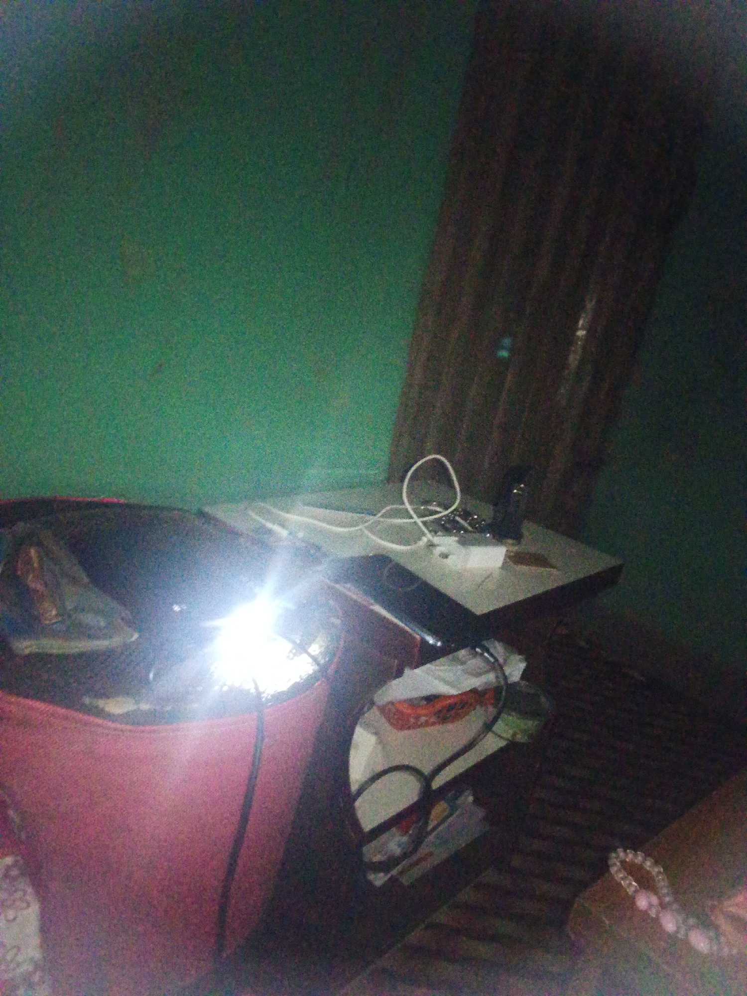 The image depicts a dimly lit room with a green wall on the left and a dark, corrugated surface on the right. In the foreground, a large, pink, cylindrical object, possibly a bin or basket, is prominently featured. A bright light source is illuminating the interior of this object, creating a glare. To the right of the pink object, a table or desk is visible, with a white electrical cord coiled on its surface, along with what appears to be a power strip and some small objects. Beneath the table, more items are present, including what looks like packaging and another black cord. On the far right, a portion of a surface with a patterned texture is visible, and a pink beaded bracelet is lying on it. The overall scene suggests an indoor environment that is cluttered and dimly lit.