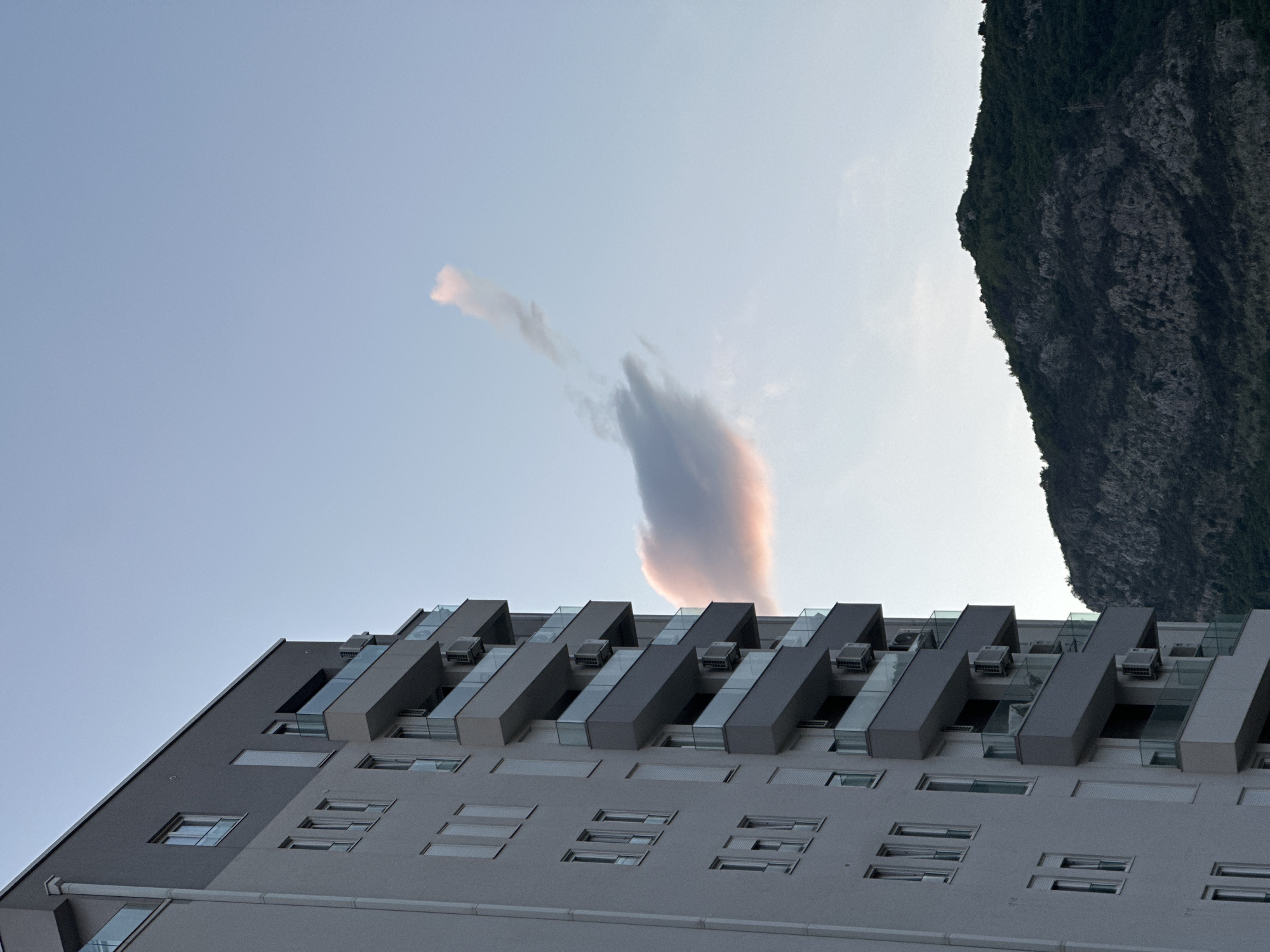 A low-angle shot shows the facade of a modern building in Budva, Montenegro, under a light blue sky with a wispy cloud formation and a portion of a mountain range. The building is predominantly gray and features rows of rectangular windows and protruding structural elements with glass railings and visible air conditioning units. The cloud formation has a vertical shape with pinkish hues near the center. The mountain range is dark green with visible rock textures. The lighting suggests either sunrise or sunset.