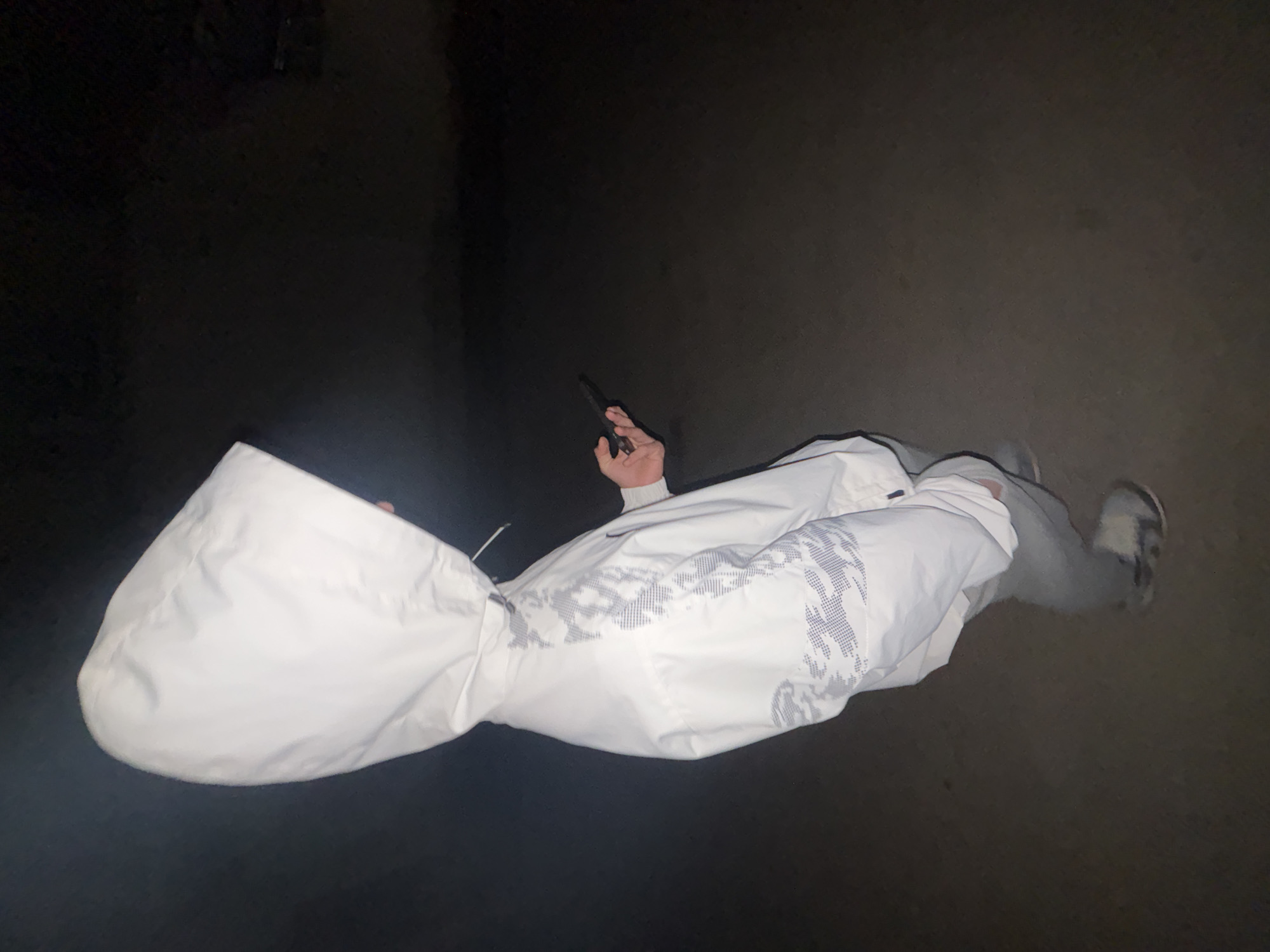 A person is standing in an unknown location. The person is wearing a white hooded jacket and light grey pants. They are holding a dark, rectangular object, possibly a phone, in their right hand. The environment is dark and indistinct, with a concrete surface underfoot. The overall lighting is dim. The exact location within the city of Lehchevo, Bulgaria is undeterminable from the image.