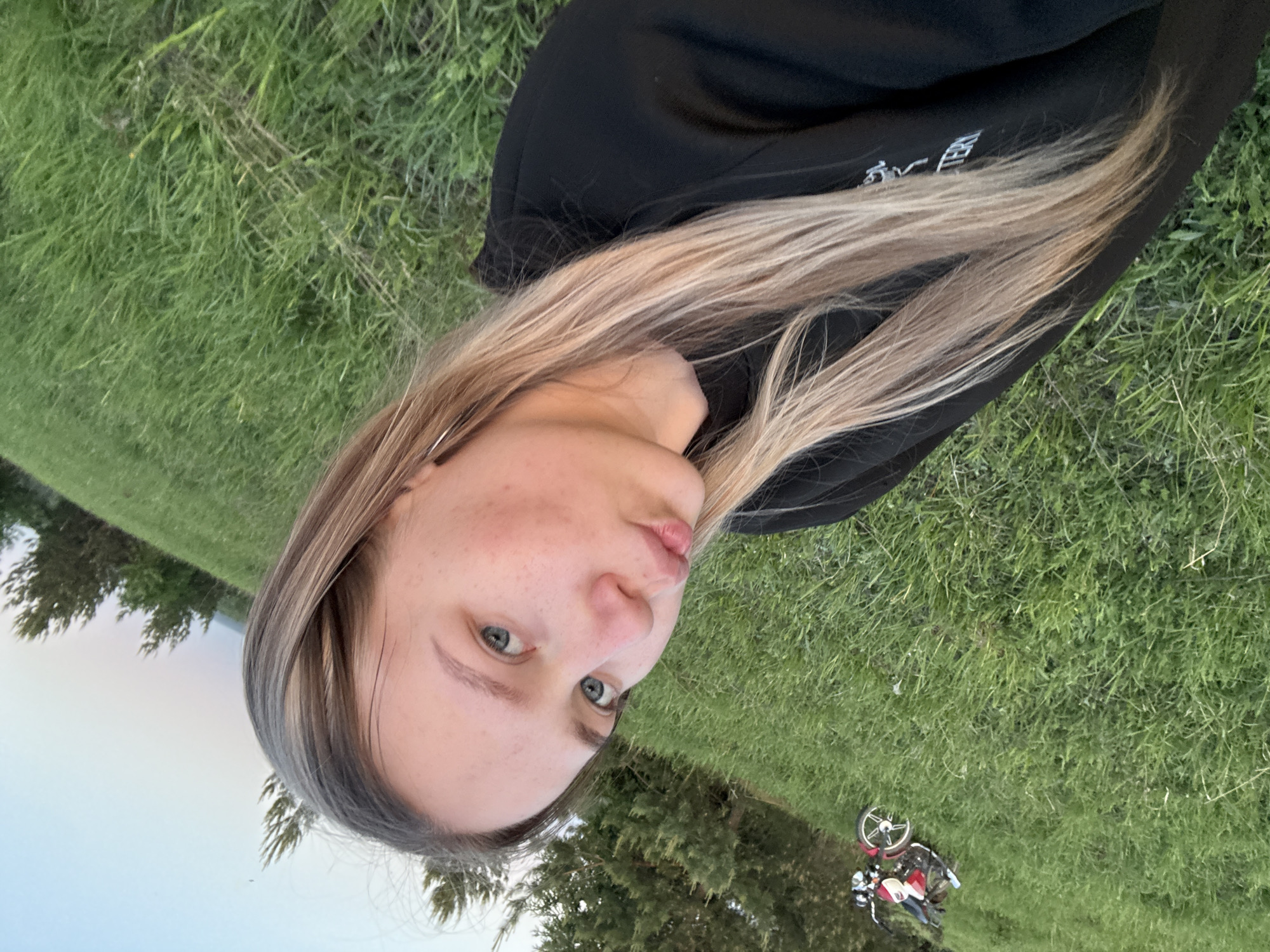A woman lies on her side in a grassy field. She has long blonde hair, and is wearing a black hooded sweatshirt. The field is located in Zhigalki, Russia. A small motorbike or scooter is visible on the grass nearby, partially obscured by trees. The sky is overcast.