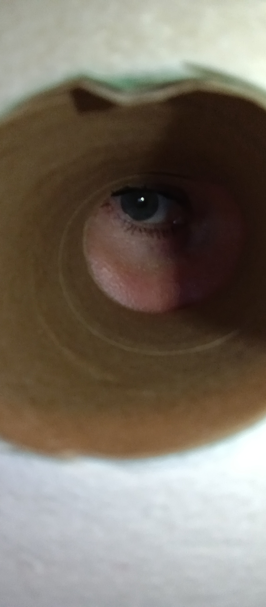 A close-up shot depicts a human eye peering through a cardboard tube. The eye is blue with a black pupil and is centered in the frame. The surrounding skin is pale and slightly reddened, particularly around the eye socket. The cardboard tube is brown and features distinct circular ridges and some imperfections along its edge. The background is a blurry white.