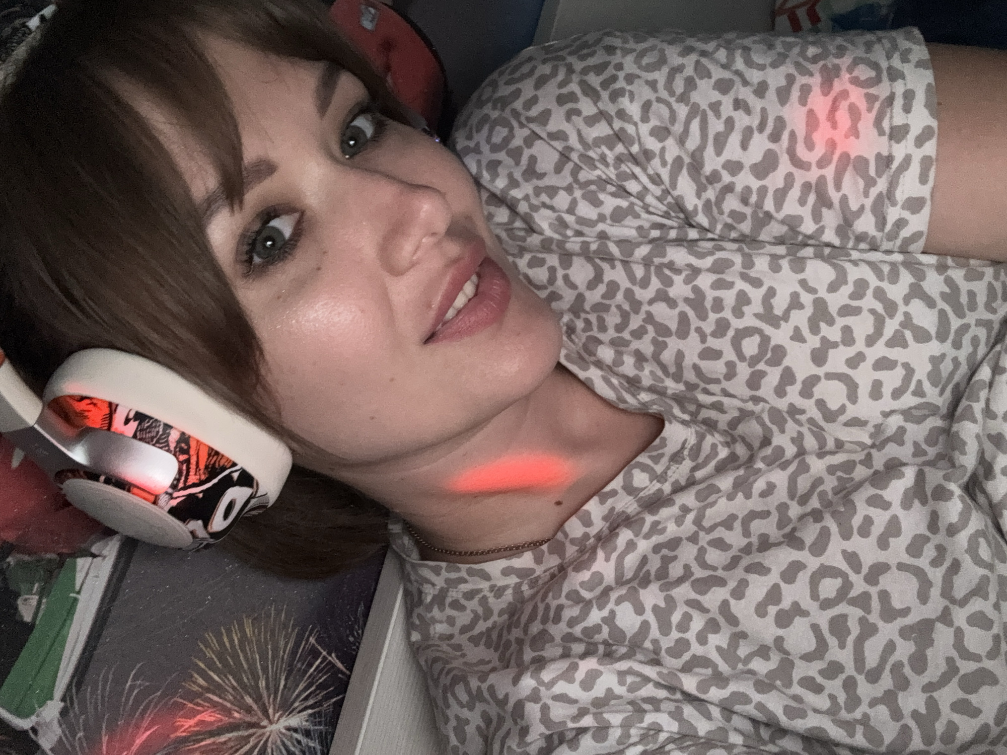 The scene is located in Chaikovsky, Russia. A single adult female is depicted in a close-up, self-portrait photograph, looking directly into the camera with a slight smile. She has brown hair with bangs, green eyes, and light-toned skin. She is wearing a light grey t-shirt with a darker grey leopard print pattern. A thin, metallic chain necklace is visible around her neck. She wears white over-ear headphones with black, white, and red graphic designs on the earcups, which emit a red light. Additional red light is evident as a vertical streak illuminating the left side of her neck and collarbone, and a diffused glow is visible on the right sleeve of her t-shirt. The background behind her left shoulder features a dark image or poster displaying red and white starbursts, resembling fireworks. A lighter, horizontal surface, possibly a shelf or headboard, is visible behind her head. The overall lighting is dim, with the red light sources providing localized illumination.