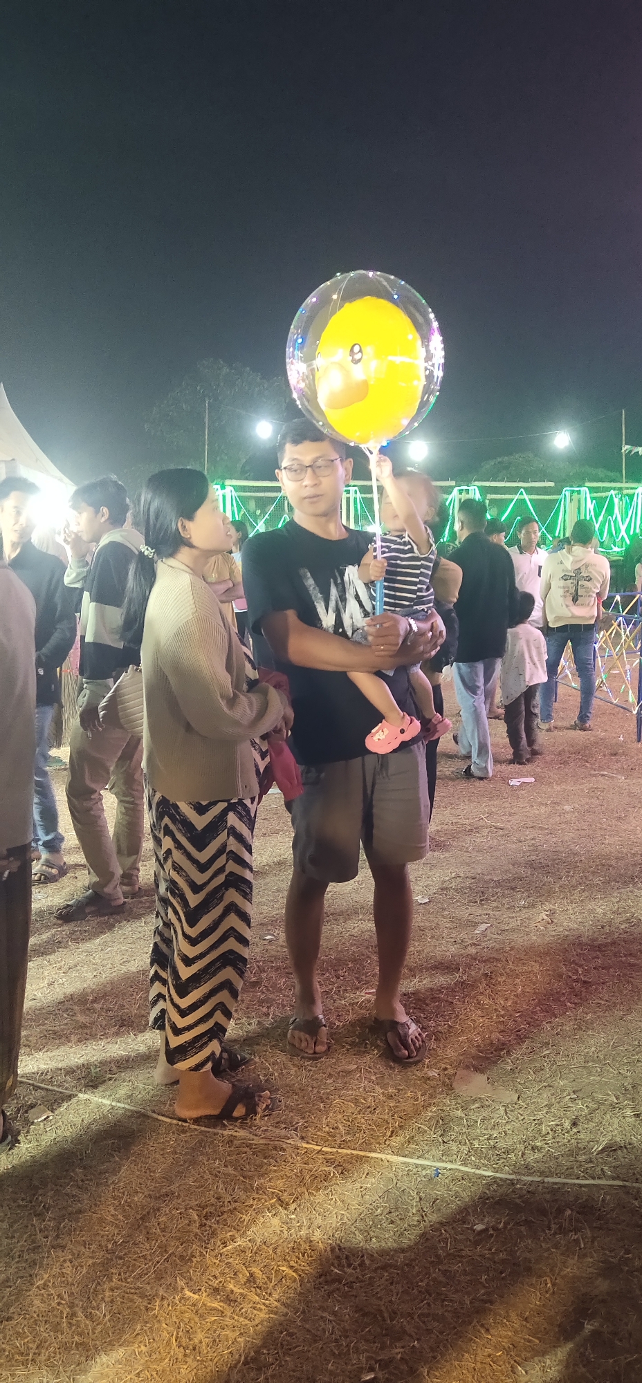 This image depicts an outdoor nighttime scene, possibly a fair or public gathering, in Tasikmadu, Indonesia. The foreground is dominated by dry grass and scattered debris, with shadows cast by artificial lighting. In the center of the frame, a man wearing glasses, a black t-shirt, and gray shorts is holding a young child. The child, dressed in a striped shirt and pink sandals, is reaching for a large, translucent yellow balloon with a duck-like face. Beside them, a woman in a beige cardigan and black-and-white chevron-patterned pants stands facing the man and child. Other individuals, mostly out of focus, are visible in the background, suggesting a crowded environment. Decorative string lights are visible in the distance, further indicating a festive event.