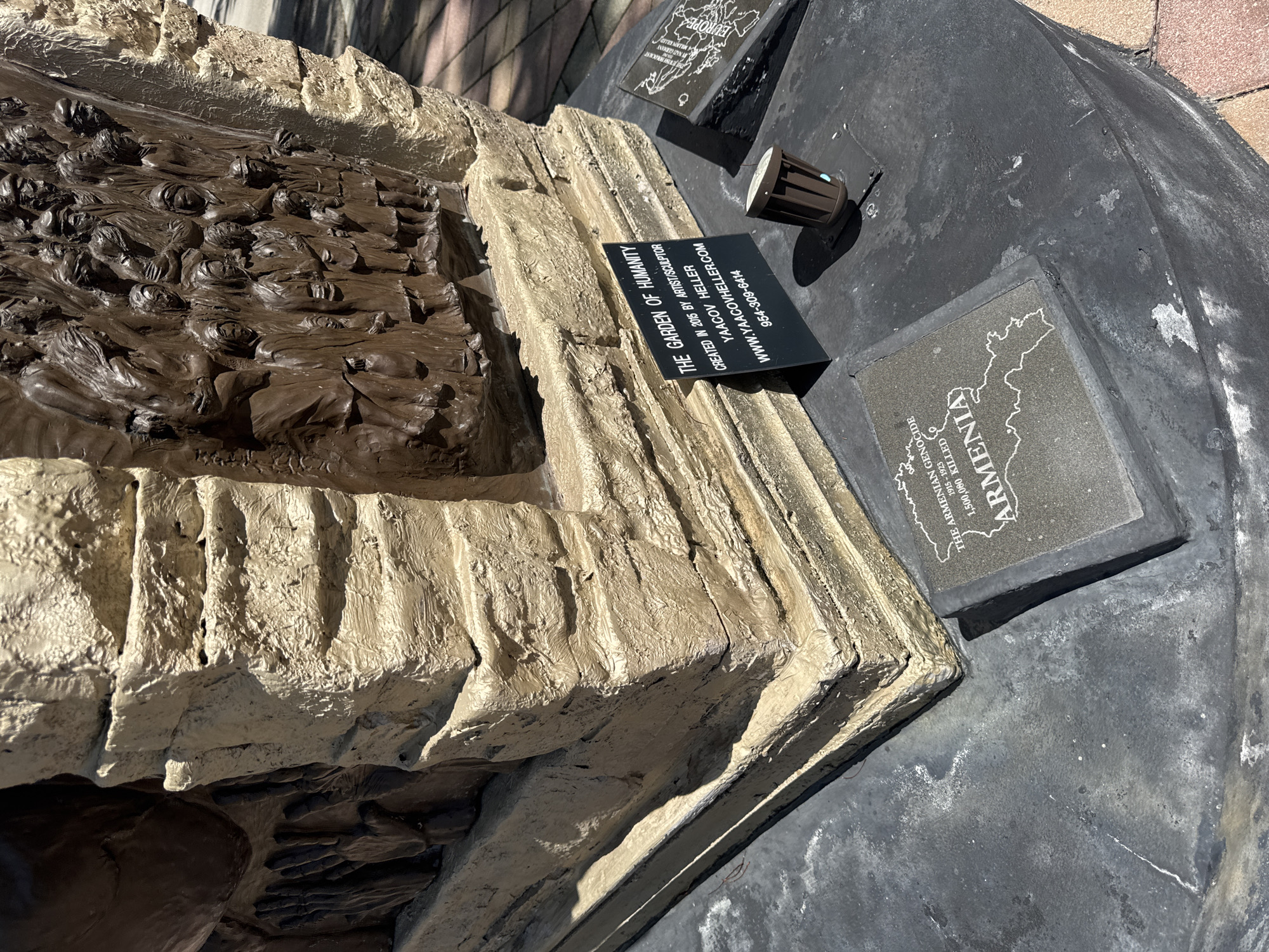 An outdoor monument is depicted from a low-angle perspective, featuring a roughly textured, multi-layered base painted a light brown or tan, mimicking stone blocks. Integrated into the monument are bronze-toned relief panels. The most prominent panel on the right displays numerous human figures in high relief, densely packed and appearing to move or gather, rendered with somber expressions and simple clothing. A smaller section of another panel on the left shows multiple hands extended from a dark background.
At the base of the monument, on a dark, coarse-textured ground surface, are three plaques. Directly in front of the sculpture is a black rectangular sign with white lettering, stating: "THE GARDEN OF HUMANITY / CREATED IN 2015 BY ARTIST/SCULPTOR / YAACOV HELLER / WWW.YAACOVHELLER.COM / 954-309-6414." To the left of this sign is a rectangular gray plaque with an etched outline map of Armenia, displaying white text: "THE ARMENIAN GENOCIDE / 1915 - 1923 / 1,500,000 KILLED / ARMENIA." A third, partially visible gray plaque on the right side also features an etched map and the word "EUROPE."
A small, cylindrical, dark brown outdoor spotlight fixture is positioned on the dark ground between the monument and the black informational plaque. The ground surface shows some white scuffs and minor debris. Beyond the dark ground, a patterned pavement composed of light-colored bricks or tiles is visible. The scene is illuminated by direct sunlight, casting distinct shadows. This monument is located in Boca Raton, United States of America.