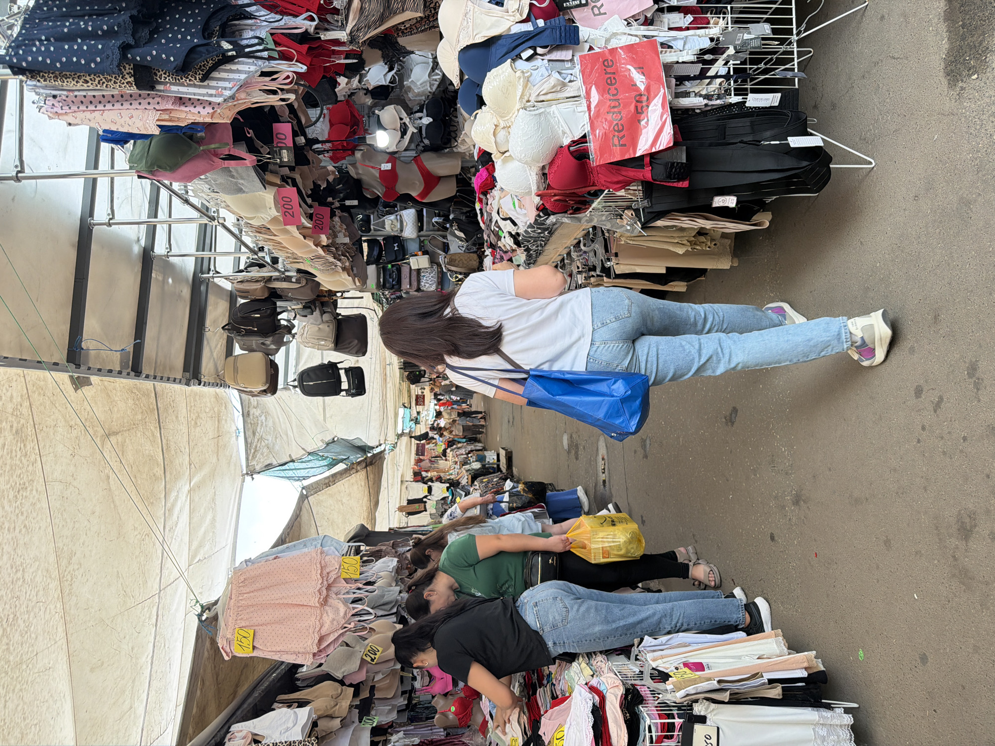 The image depicts an outdoor marketplace, likely a street market, in Chișinău, Moldova, with stalls selling undergarments and clothing. In the foreground, a woman with dark hair, wearing a white t-shirt and light blue jeans, walks away from the camera, carrying a bright blue tote bag. To her left, two women are browsing at a stall. The woman closer to the camera wears a black t-shirt and light blue jeans, while the woman next to her is in a green t-shirt and dark pants, holding a yellow plastic bag. The stalls are covered by beige tarpaulins stretched between metal poles. The merchandise, primarily bras and lingerie, is displayed on hangers and shelves. Red price tags are visible, one with "Reducere" and "150 lei" printed on it. Other shoppers and stalls are visible in the background, indicating a busy market environment. The ground is paved with asphalt.