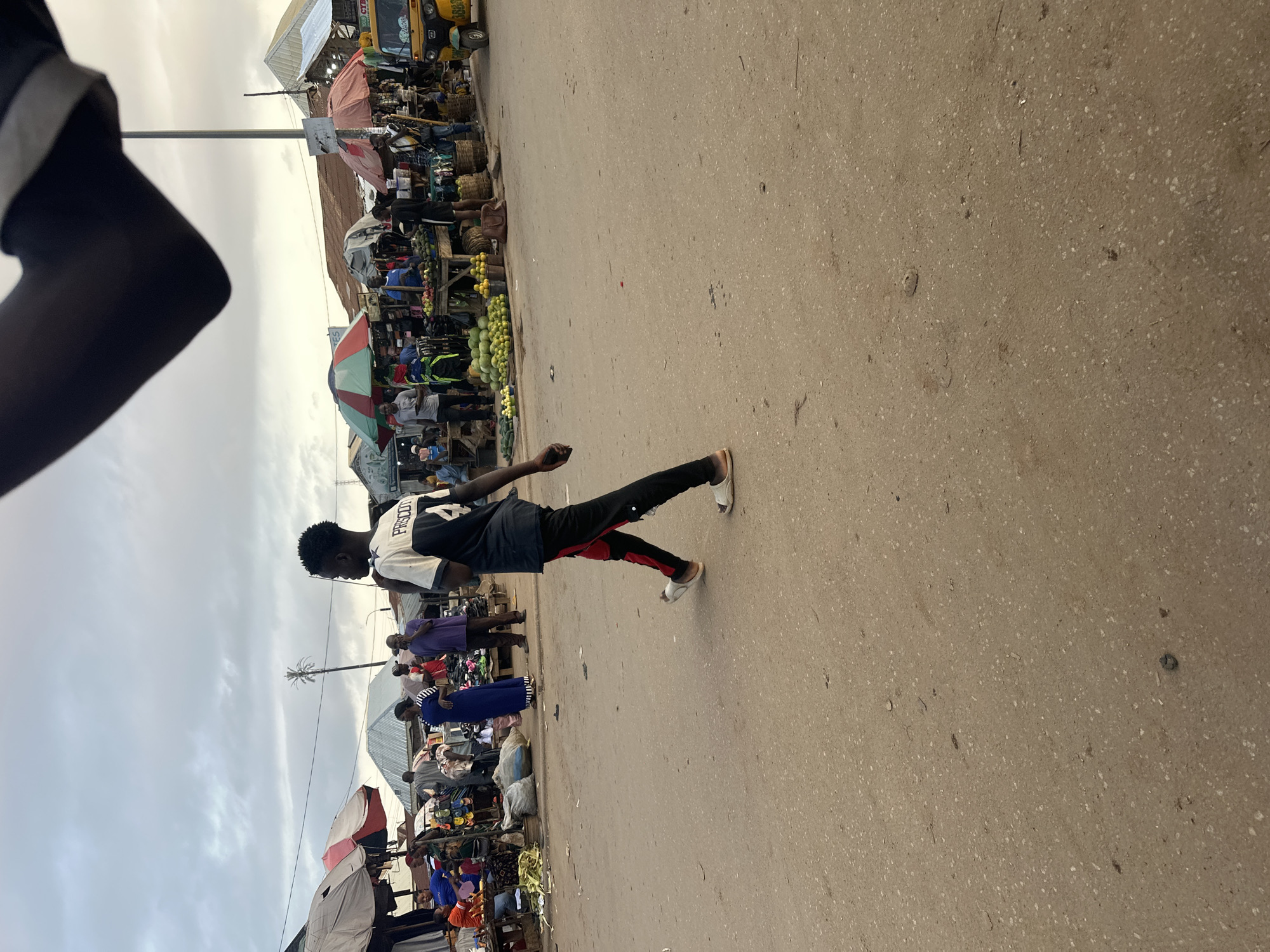 The image depicts a bustling outdoor market scene, likely in Abuja, Nigeria. The primary subject is a young man walking across a paved area, seen from the side. He is wearing a black t-shirt with white lettering that reads "FRISCOT" and has red and black striped pants. His stance suggests he is in motion.
Behind him, the market is crowded with people. Stalls are set up along a street, displaying various goods, including what appear to be fruits and vegetables, particularly a large pile of green produce, possibly watermelons or melons. There are umbrellas providing shade over some of the stalls. People are milling about, browsing and interacting, creating a sense of activity and commerce. A yellow vehicle, possibly a bus or truck, is visible in the background, suggesting transportation is part of the scene.
The time of day appears to be daytime, given the lighting, though the sky is overcast with some clouds, indicating potentially dull or pre-rain weather. The ground is a sandy-colored pavement, typical of outdoor markets. The overall atmosphere conveyed is one of lively daily life and trade within a busy urban or semi-urban environment. No specific emotions are clearly discernible, but the presence of many people engaged in shopping and selling implies a functional and active community hub.
