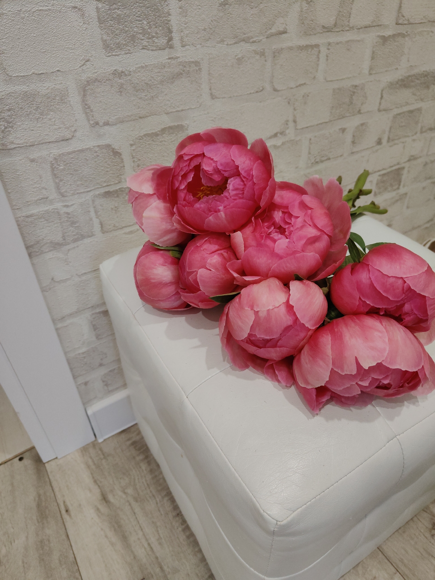 A cluster of approximately eight vibrant pink peonies, some in partial bloom and others fully open, is positioned centrally on a white, square, quilted ottoman. The peonies display layered petals and visible yellow stamens. The ottoman is upholstered in a material resembling leather, featuring a stitched, quilted pattern on its top and sides. The background consists of a wall finished with a light-colored, textured surface mimicking off-white or light grey bricks with darker grout lines. To the left, a vertical white architectural trim, possibly a door frame, extends downwards to meet a white baseboard. The floor below the ottoman and baseboard is composed of light-toned, horizontally oriented wooden or laminate planks, exhibiting a visible grain. The scene is illuminated by ambient indoor light. No individuals or animals are present. The location is in the city of Unknown, Unknown.