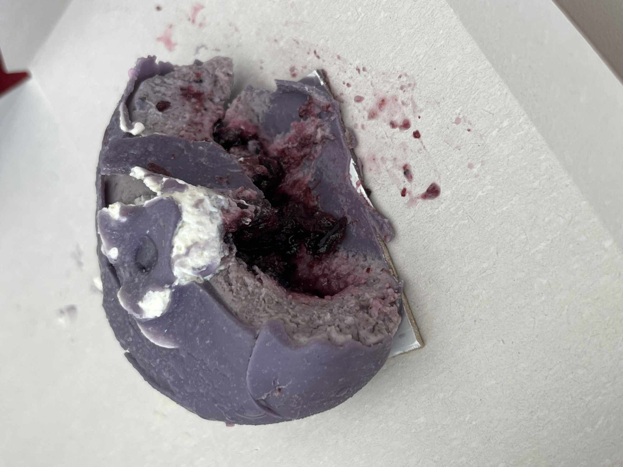 The image presents a close-up view of a partially consumed or broken dessert item resting on a light-colored, textured surface, likely a paperboard container or tray. The dessert is predominantly purple in color, suggesting flavors like ube, taro, or a berry-based ingredient in its outer layer, which appears to be a soft cake, pastry, or mousse.
A significant portion of the dessert has been scooped out or broken, exposing a dark red to deep purple filling at its center. This filling contains visible pieces of fruit, resembling berries such as blueberries or raspberries, and their rich juices have stained the surrounding inner layers of the dessert. Several dollops and streaks of white cream or frosting are visible on the purple exterior and within the exposed sections.
The light-colored surface around the dessert is marked with numerous splatters of the dark berry juice, indicating that the item has been actively eaten or disturbed. The texture of the purple dessert is somewhat crumbly and soft where it is broken, while the intact outer sections appear smoother. The lighting in the image is soft and even, highlighting the colors and textures of the food. There are no people, explicit location cues, or discernible text visible in the frame. The scene captures the aftermath of enjoying a fruity, creamy dessert.