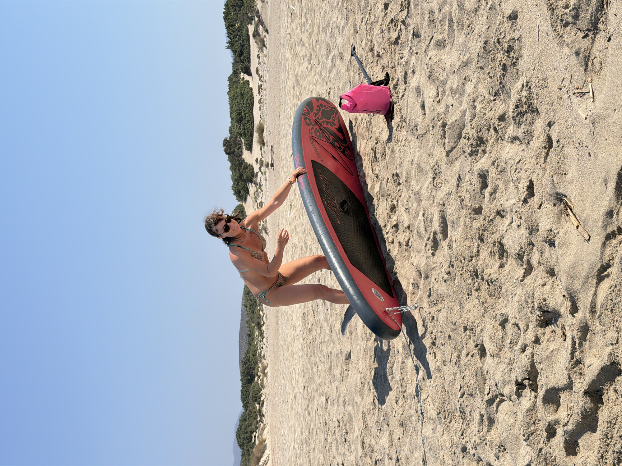 A woman in a bikini kneels on a sandy beach, reaching for a red and black stand-up paddleboard. She is wearing sunglasses and her dark hair is slightly windswept. Beside the paddleboard, to its right, is a bright pink dry bag and a black paddle. The background consists of sand dunes covered in scrubby green vegetation, with a clear blue sky above. The scene appears to be in Greece, though the specific city is unknown.