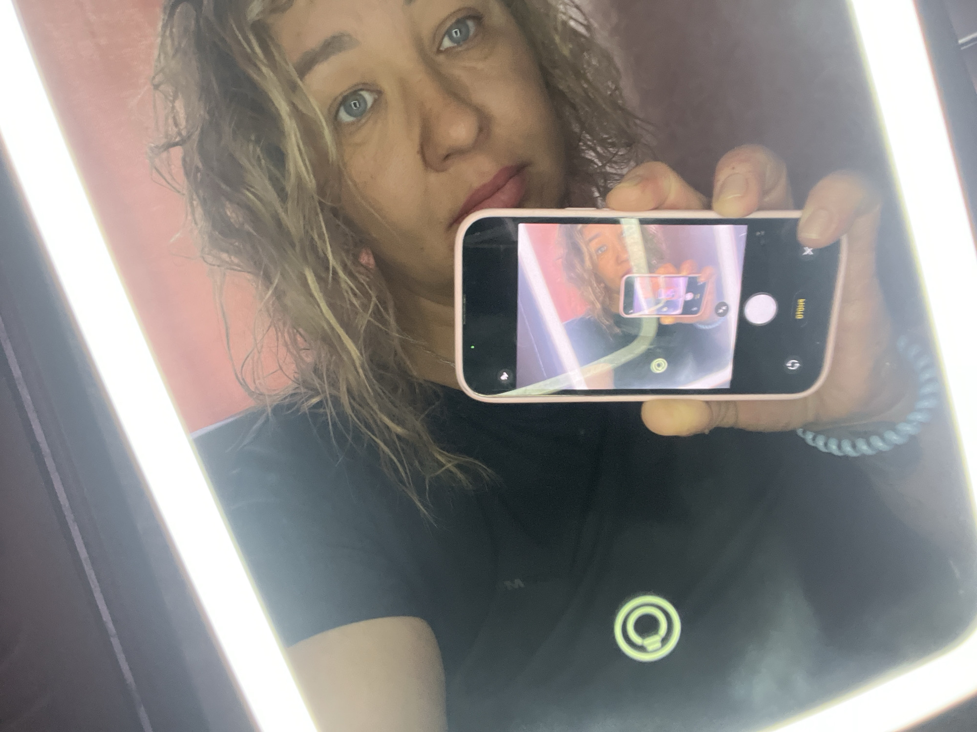 A woman with blonde, wavy hair and blue eyes takes a selfie in a mirror. She is wearing a black t-shirt. She holds a pink smartphone in her right hand, which is capturing her reflection. The background appears to be a room with a bright, vertical light source on the left and a red-toned wall behind her. The image is taken from a close-up perspective, focusing on the woman's face and the phone. There is no discernible indication of the city of Chaikovsky, Russia within the image.