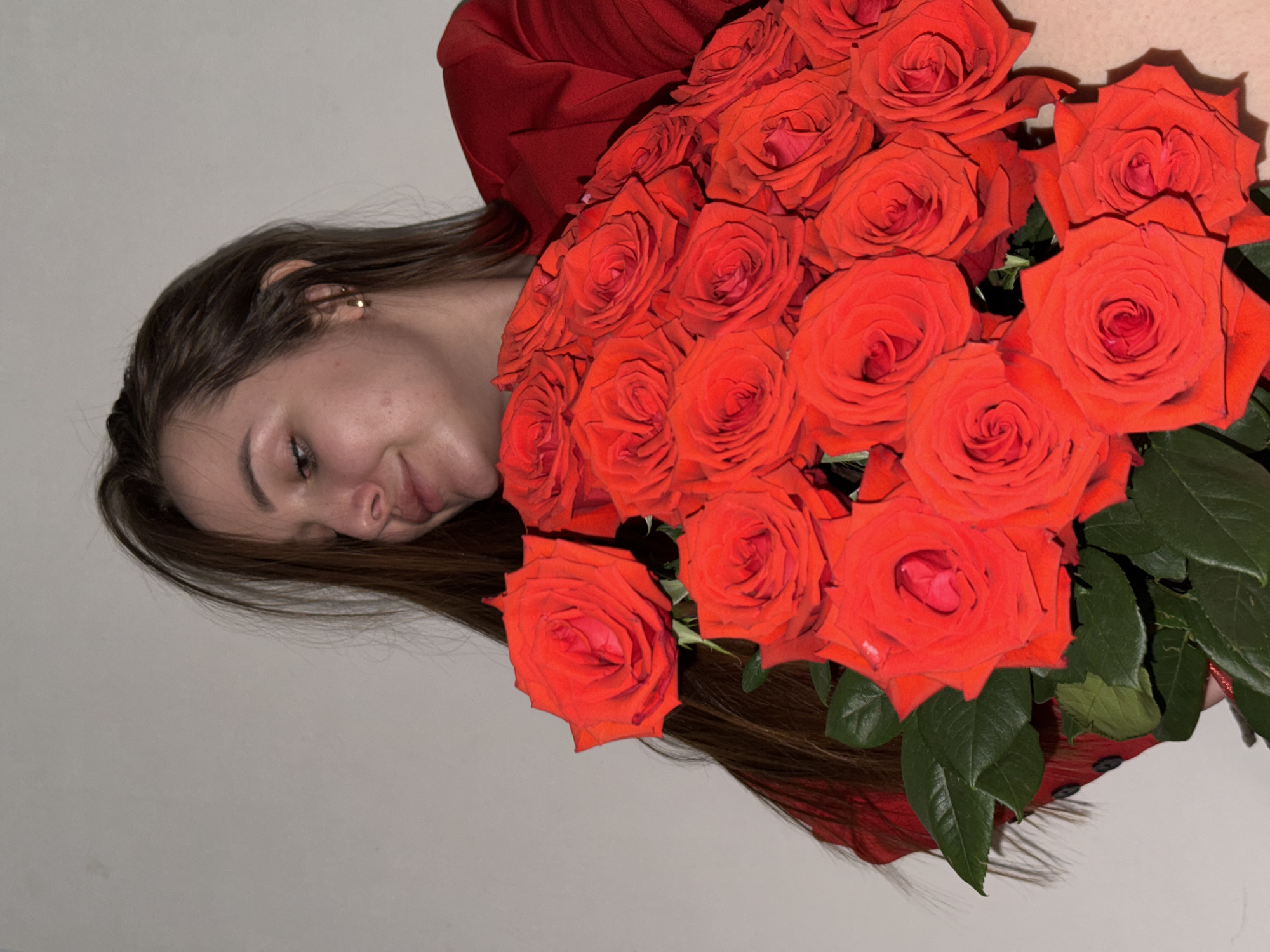 A woman is holding a large bouquet of red roses close to her face. The woman has brown hair and a red top. The roses are a vibrant red and the bouquet is dense with blossoms. The background is a plain, light color. The location in the city of Chaikovsky, Russia is unknown.