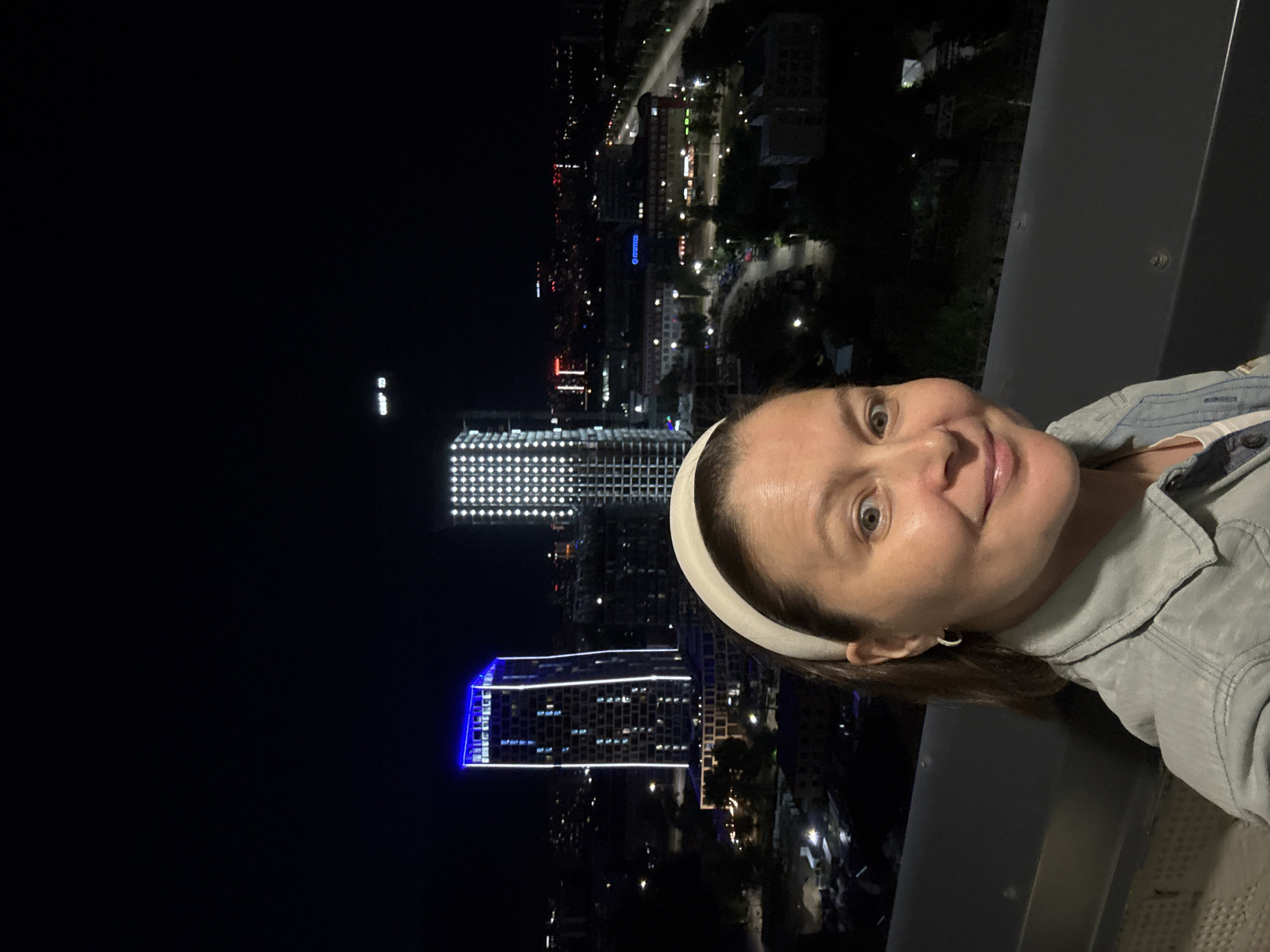 A woman with brown hair, wearing a white headband, and a light denim jacket takes a selfie at night with a cityscape in the background. She has a light smile and is looking directly at the camera. The background shows several illuminated buildings, including a tall skyscraper with blue and white lights on its facade. Another building to the right is illuminated with many small white lights. The city lights extend into the distance, suggesting an urban environment. The photo appears to be taken from an elevated position, possibly a balcony or rooftop. The city in the background is Izhevsk, Russia, as indicated by the presence of the building labeled "GAZPROM NEFT 70".