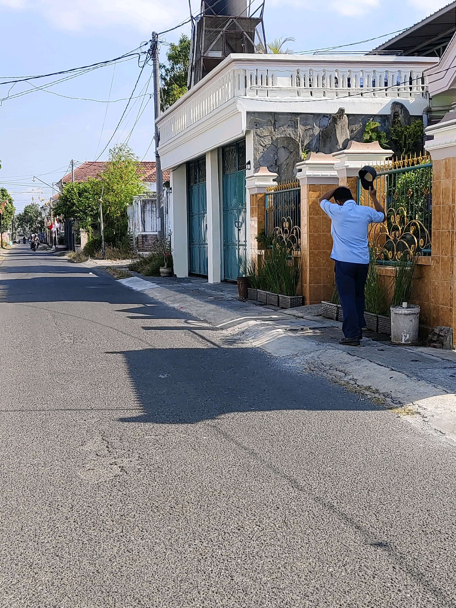 In Probolinggo, Indonesia, a man in a light blue short-sleeved shirt and dark pants stands on the side of a paved street, holding a dark cap in his right hand. He appears to be adjusting his hair or hat. The street is lined with houses and vegetation. To the right, a decorative gold fence with a patterned design encloses a property with orange tiled walls. Behind the fence, a white building with a white balustrade on the second level is visible, featuring stone-like cladding on the lower part of its facade. Several long, thin planters with green grass-like plants are placed along the fence. A cylindrical trash receptacle stands to the right of the fence. Overhead, power lines crisscross the sky. The scene is illuminated by daylight, casting shadows on the street.