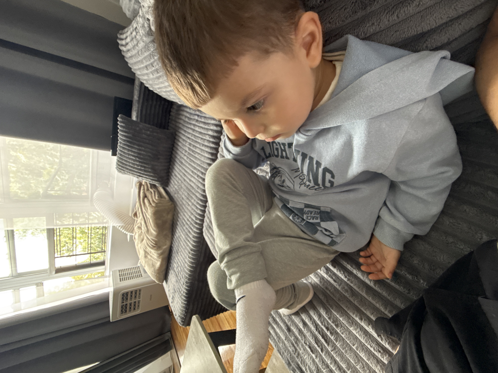 A young child, appearing to be male and of Caucasian descent, is reclining on a grey couch in a room that appears to be a living area. The child is wearing a light blue hooded sweatshirt with checkered detailing and the words "RACE READY" printed on it, along with grey sweatpants and white socks. The child's legs are crossed, and their right arm is resting on their chest while their left hand is near their face. The couch has a textured fabric and is adorned with several grey throw pillows and a folded tan blanket.
In the background, a window with white blinds and grey curtains is visible, offering a view of green foliage outside. To the left of the window, a white portable air conditioner unit is positioned on the floor, with a flexible hose extending towards the window. A grey coffee table is situated to the left of the couch. The overall impression is that of a comfortable indoor setting, possibly a home in New York City, United States of America, given the common urban apartment elements like portable AC units and window views.