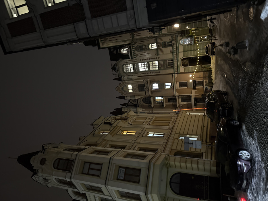 A nighttime street scene with ornate, illuminated buildings lining either side. Parked cars are visible along the right side of the street, with some snow or ice present. The sky is dark, suggesting it's late at night. The buildings exhibit varied architectural styles, featuring multiple windows and decorative elements. Streetlights and lighting within the buildings cast a warm glow.