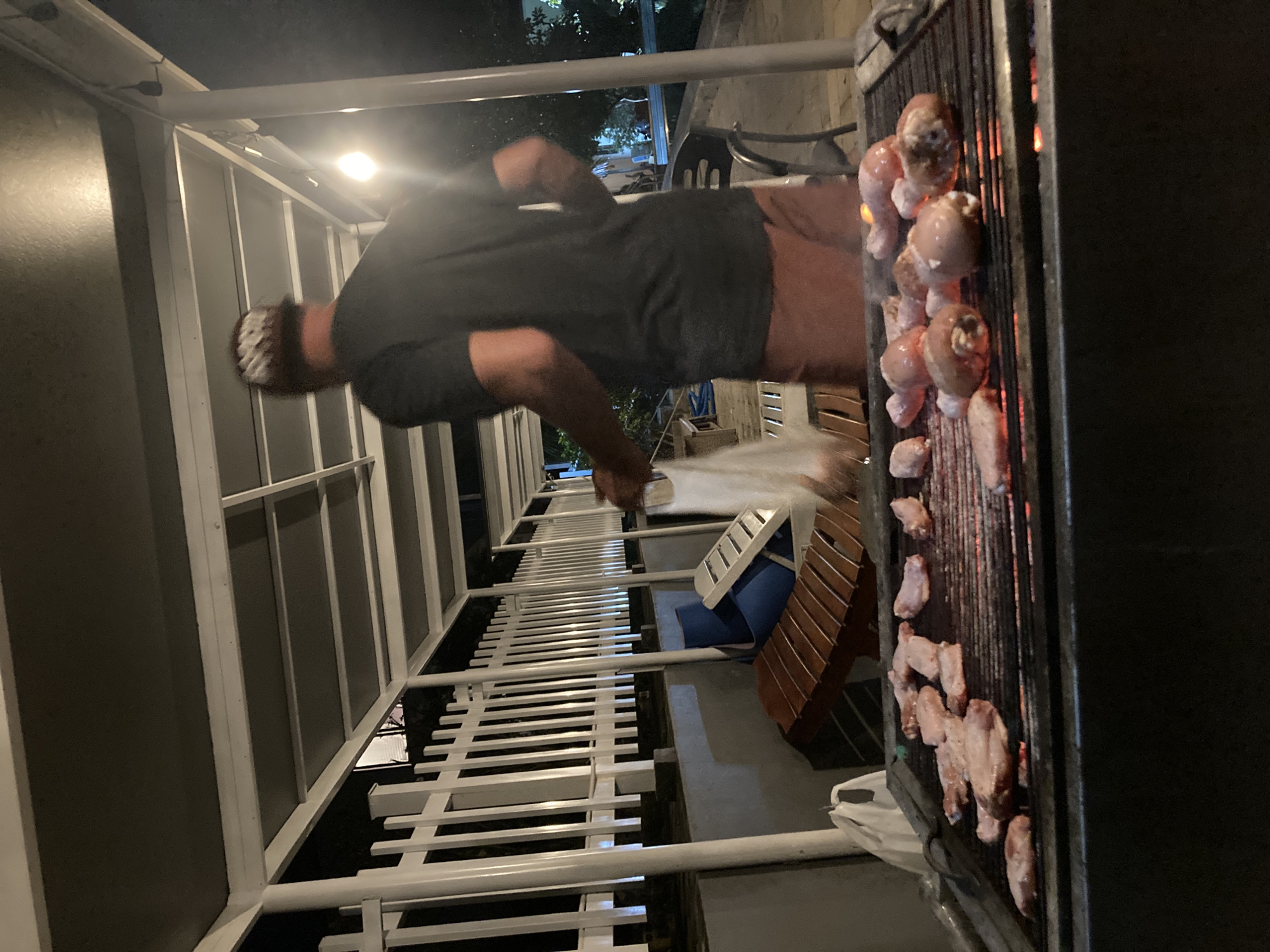A man is grilling meat on a grill on a balcony. The balcony has white railings and a privacy screen. The meat on the grill appears to be chicken wings and drumsticks. The grill is black, and smoke is rising from the cooking meat. There is a blue and white chair and a brown stool on the balcony. The location is Mui Ne, Vietnam, but the evidence for this is not contained in the image.