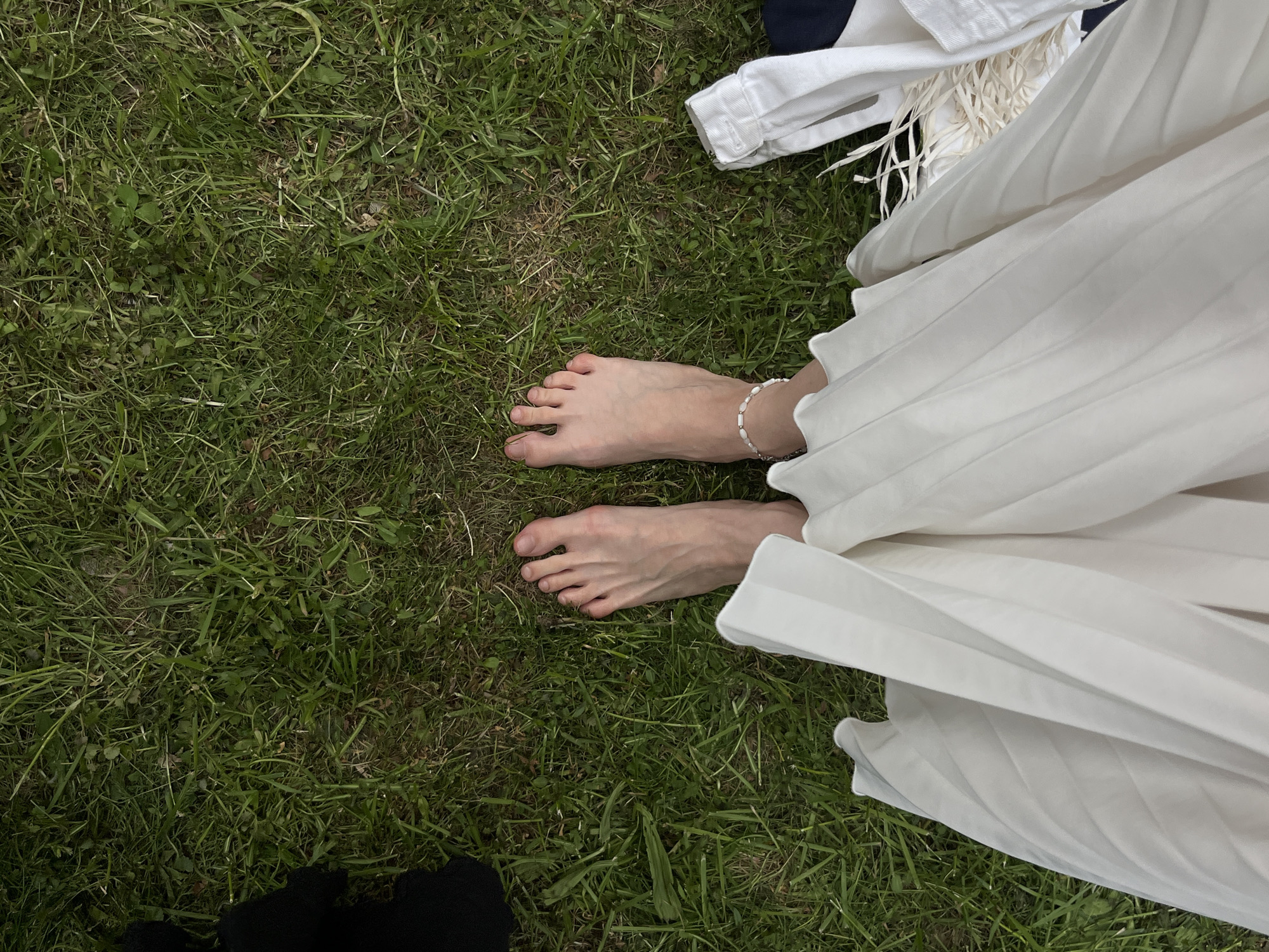 The image depicts a pair of bare feet standing on a patch of green grass. An ankle bracelet adorns one ankle. The bottom portion of a white pleated dress is visible on the right side of the frame. A white shirt and possibly a navy garment are visible in the upper right corner. The background is simply the green grass, and there is no indication that this image was taken in Moscow, Russia. It could be any location with grass.