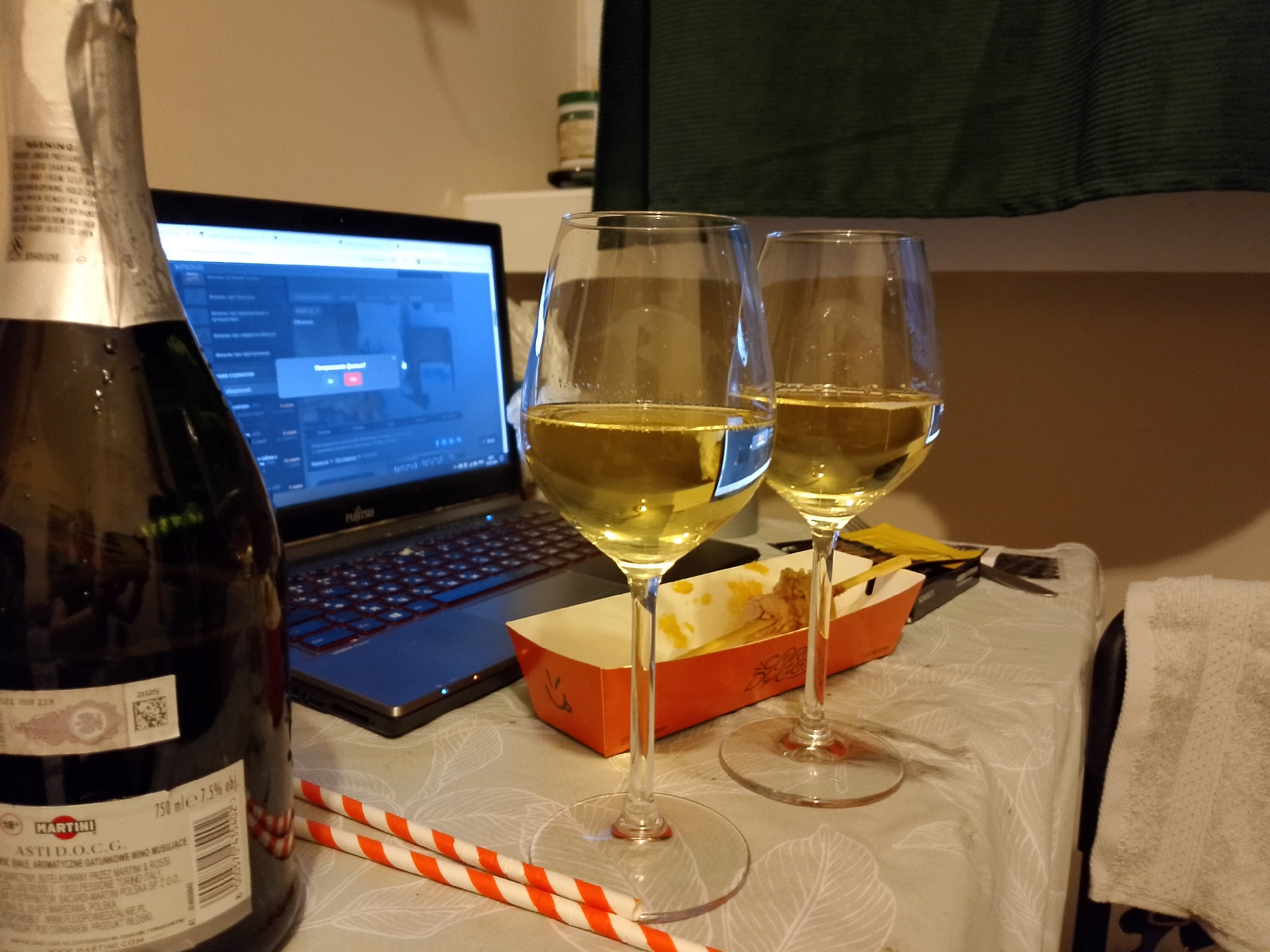 A bottle of Martini Asti D.O.C.G. sparkling wine stands on a patterned tablecloth next to an open laptop, two glasses of white wine, and a small orange carton containing food. Two orange and white striped straws are placed in front of the glasses. A white towel is draped over the edge of a chair on the right side of the frame. The scene appears to be set indoors, possibly in a home or hotel room, given the casual arrangement of items and the presence of a laptop and a towel. The lighting suggests it is nighttime or a dimly lit room.