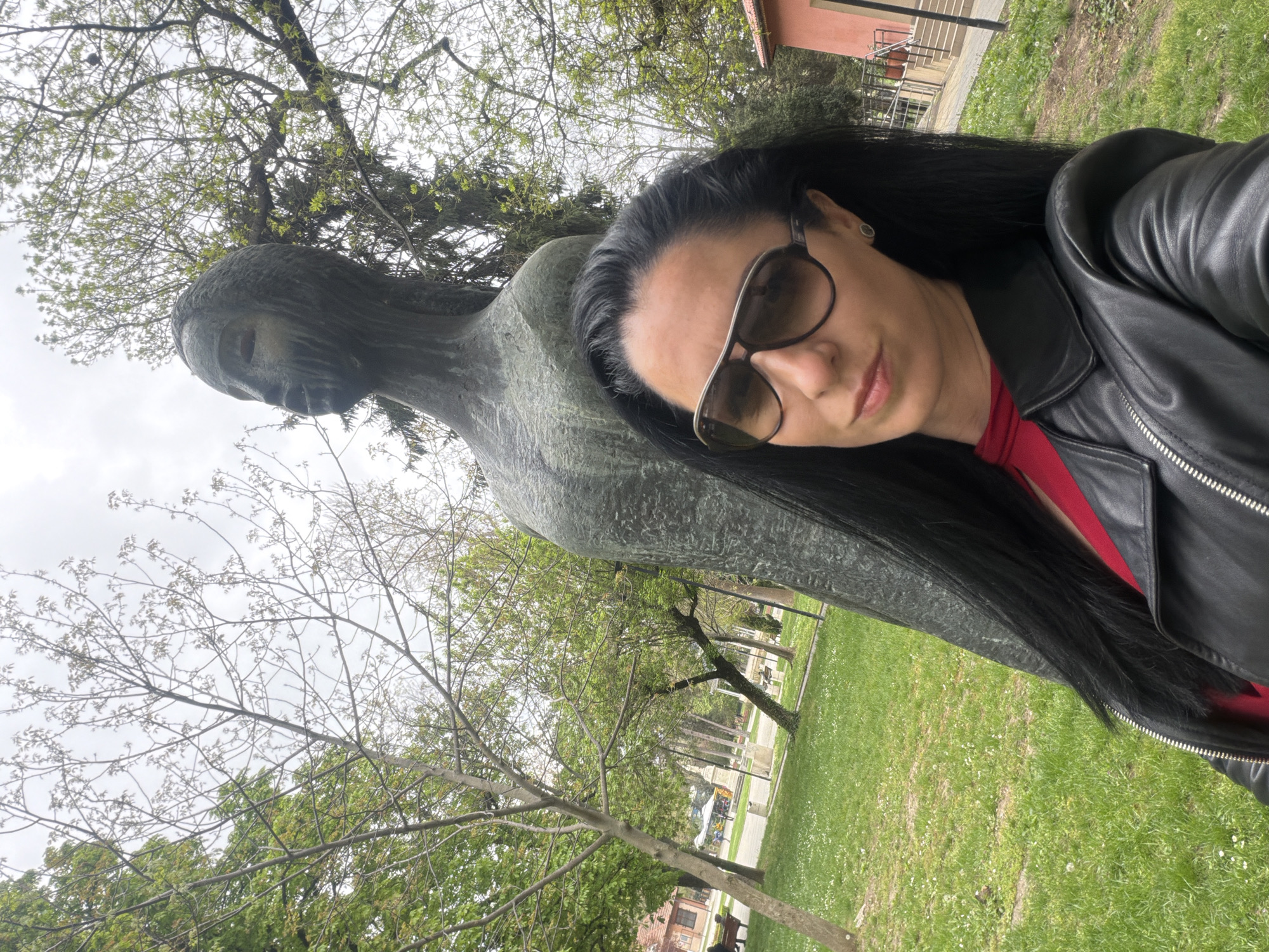 The image is a selfie taken outdoors on a grassy lawn with bare trees and budding trees visible in the background. A dark-haired woman wearing sunglasses and a black leather jacket poses next to a bronze sculpture of a figure with downcast eyes. In the background, there is a building, a paved path, and other trees, suggestive of a park setting. This is likely taken in the city of Pleven, Bulgaria, given the context of the other images.