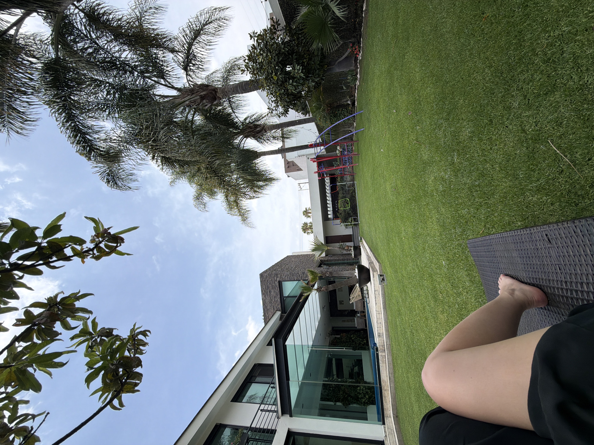The image is taken outdoors, likely from a relaxed, seated position, with a view encompassing a contemporary house, manicured lawn, and clear sky. A leg and partial view of a person wearing black clothing are visible in the foreground. The house features modern architectural elements, including expansive glass walls and sharp angles. A vibrant green lawn occupies the central space of the image, adjacent to the house. In the distance, a children's playground with red and blue structures is discernible. Trees, including palms, dot the landscape, adding to the outdoor setting. The location is in the city of Unknown, Unknown.