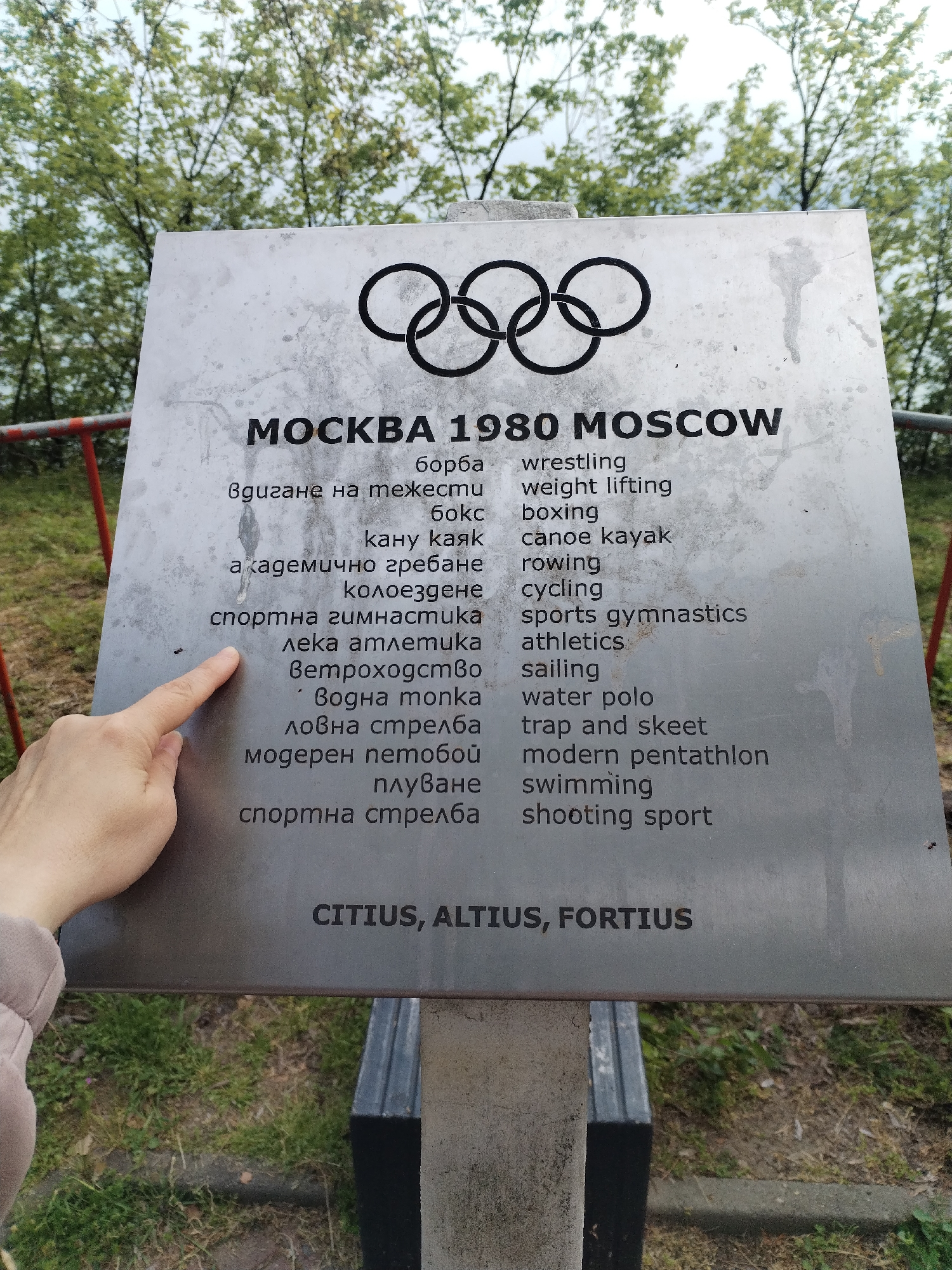 A medium-angle shot shows a rectangular metal sign commemorating the 1980 Moscow Olympics. The sign has the Olympic rings at the top, followed by "МОСКВА 1980 MOSCOW" in bold. Underneath, a list of sports is translated from Bulgarian to English. A light-skinned human hand with trimmed fingernails points towards the center-left of the sign, around the line "лека атлетика." The sign is mounted on a dark metal stand, situated outdoors. The background features green grass, some red metal railing, and trees under a cloudy sky. Given the text of the sign in both English and Bulgarian, it is likely the sign is located in a city such as Varna, Bulgaria.