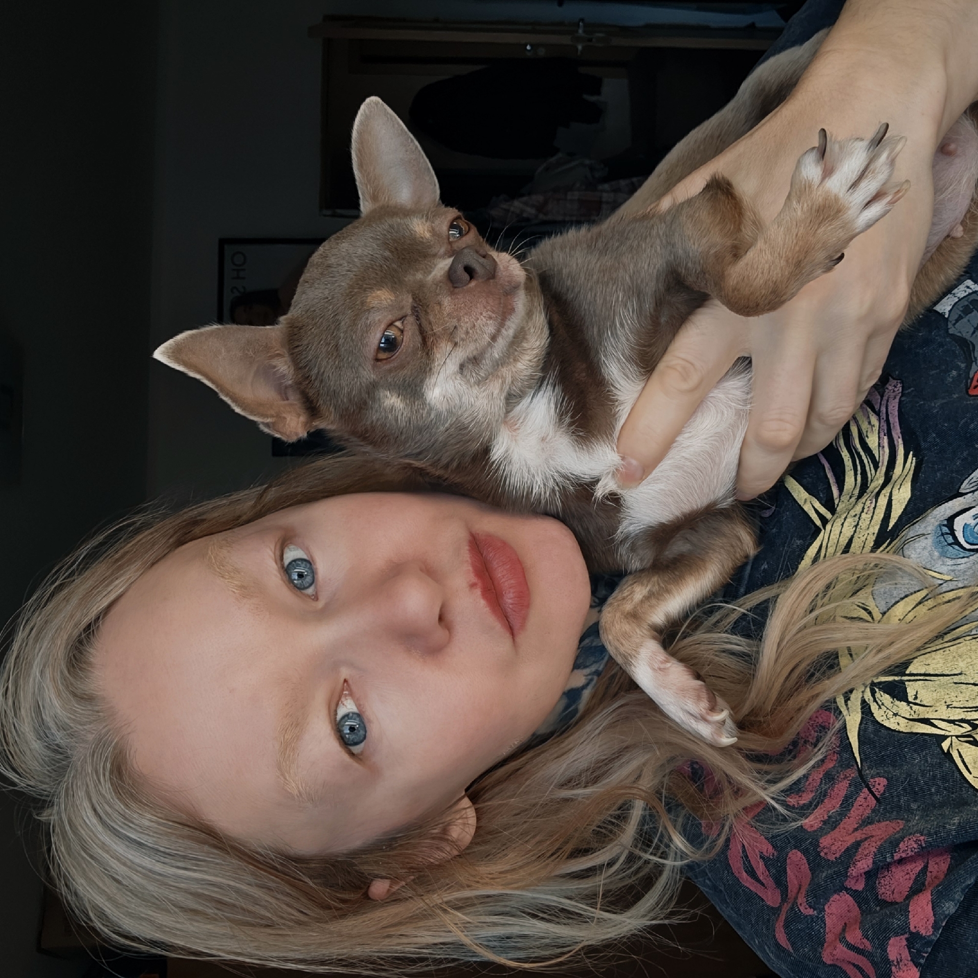 A woman with blonde hair and blue eyes is holding a brown and white chihuahua. The woman is wearing a dark blue t-shirt with a colorful design. The photo is taken indoors. It is not possible to identify the specific location as St. Petersburg, Russia based solely on the visual information.