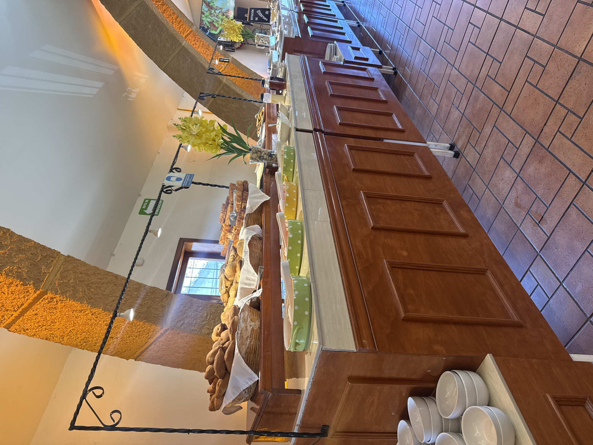 The image shows a buffet in a room with tan stone pillars and a red brick floor. The buffet table is made of dark wood with recessed paneling. Various food items, including bread, are displayed on the buffet table. There is a floral arrangement with yellow flowers on the table. The room is well-lit with natural light coming from a window and overhead lighting. White bowls are stacked at the end of the buffet. Signage is visible above the food area. Based on the architectural style and the signage, it is plausible the location is in San Cristóbal, Mexico, but there is no definitive proof from the image alone. There are no people visible in the image.