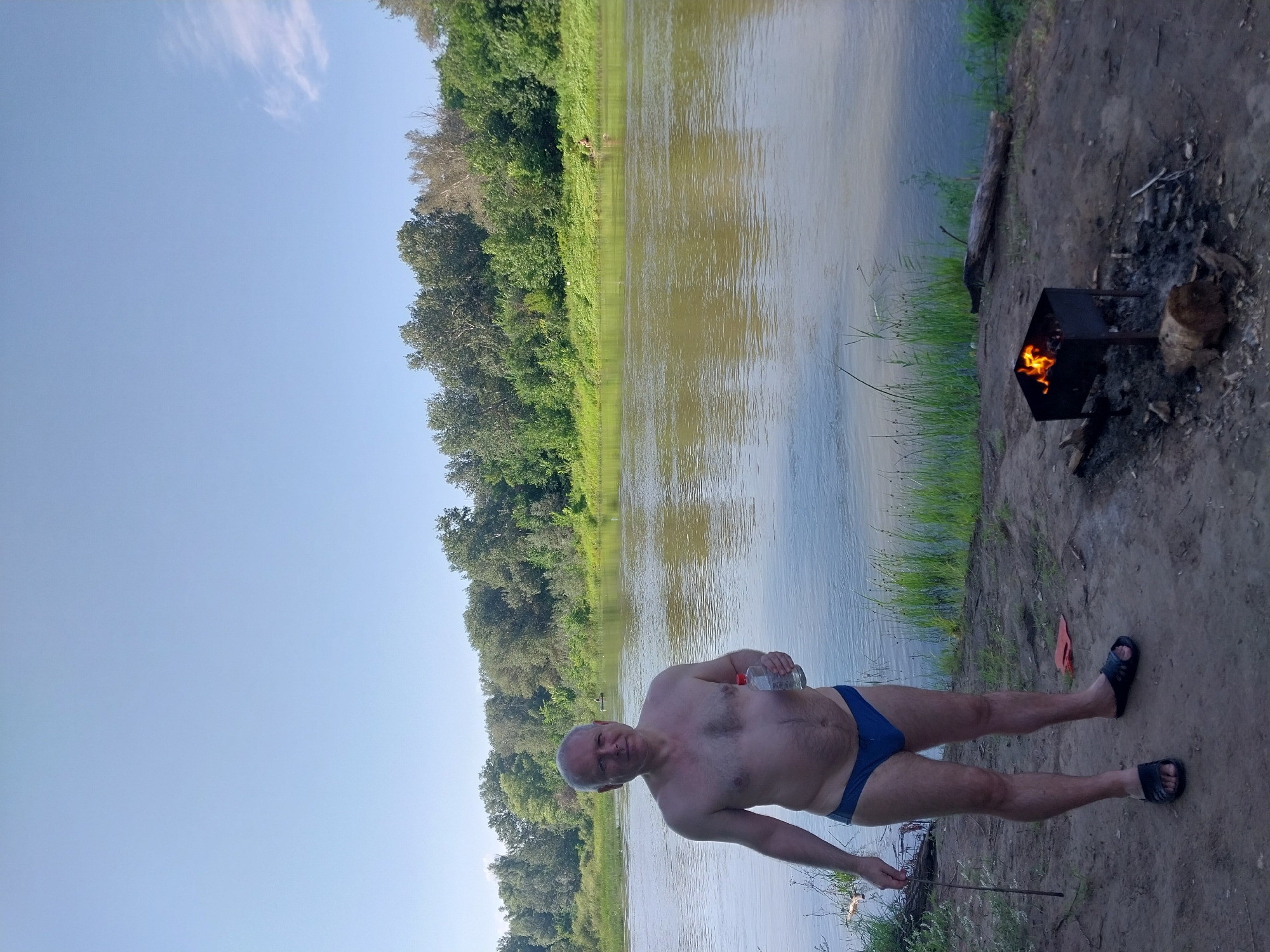 The image shows an adult male standing on the bank of a river, holding a clear plastic bottle with a white cap. He is wearing blue swimming trunks and black sandals. In the foreground, to the right of the man, is a charcoal grill with flames burning inside. There is also a log and some burnt wood on the ground near the grill. The river is wide and calm, with a tree line on the opposite bank. The sky is clear and blue. The location is the bank of the Samara River in Chkalov, Russia. The man appears to be preparing for or enjoying a barbecue by the river.
