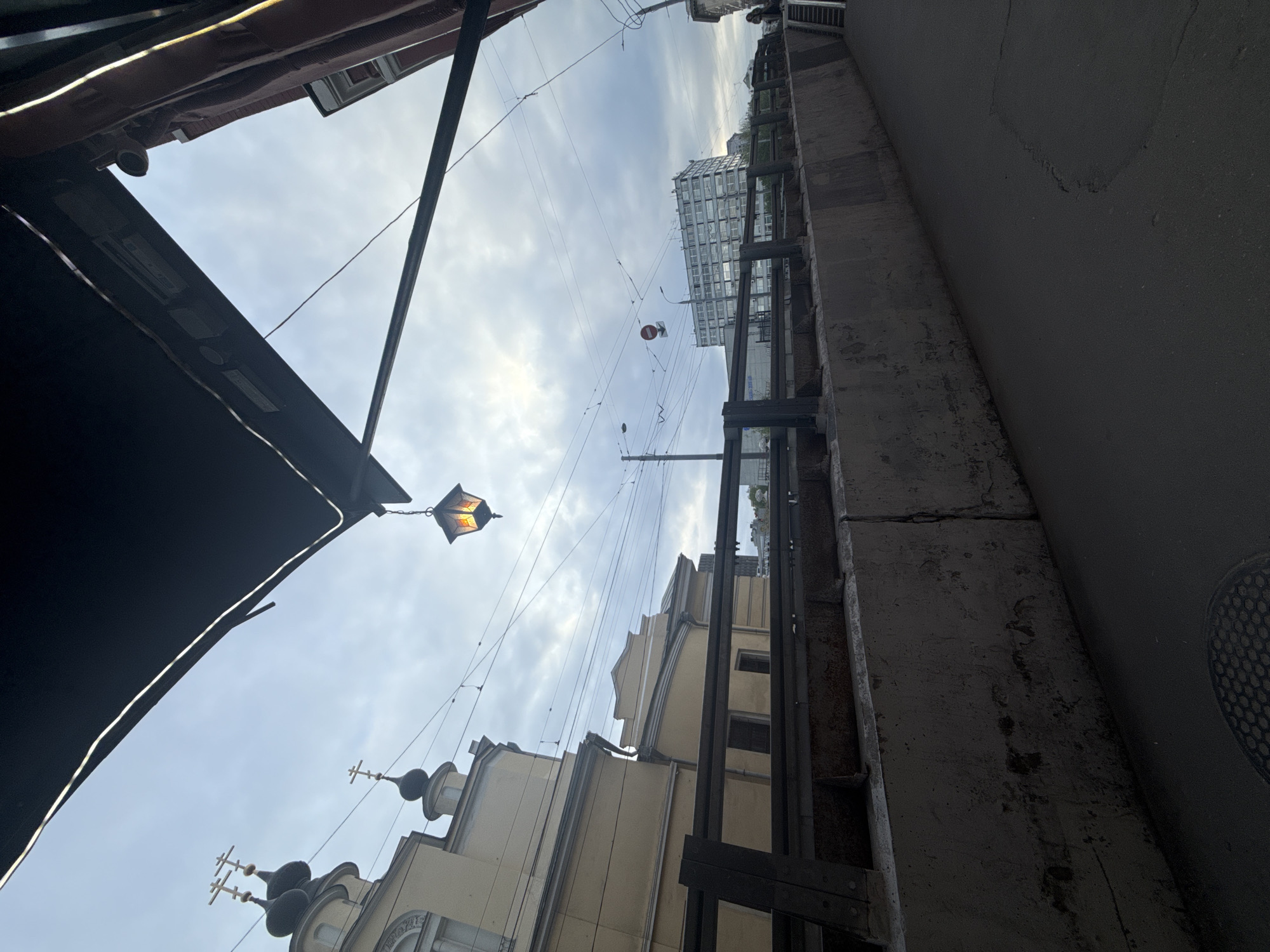 The image presents a view from street level, looking upwards, revealing a narrow urban canyon. On the left, a dark, angular awning partially obscures the sky. A decorative street lamp, lit, hangs suspended from above. Dominating the scene are buildings with varied architectural styles. To the right, a concrete wall stretches upwards. A building with crosses on top is visible. Overhead, multiple electrical wires crisscross the sky. A modern building is visible in the distance. The location is Moscow, Russia.