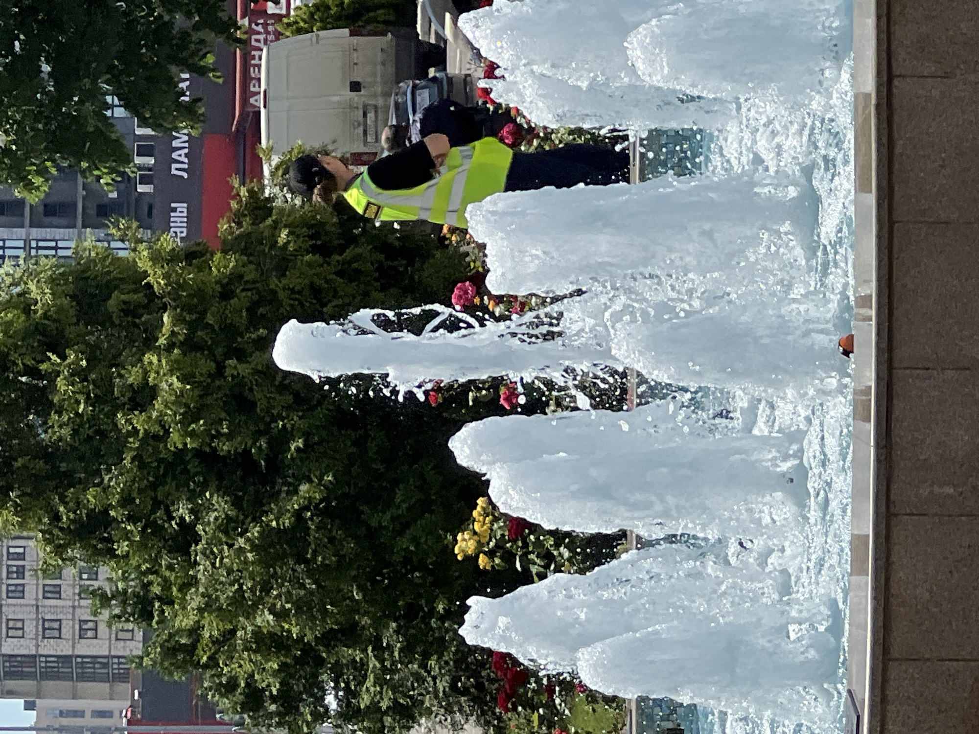 The image captures an outdoor scene featuring a vibrant fountain, lush greenery, and a person in a high-visibility vest. In the foreground and extending to the right, a multi-jet fountain actively sprays tall columns of white water, creating a refreshing atmosphere. The fountain basin edge, made of light-colored stone or concrete, is visible on the far right.
To the left of the fountain, a person is standing, partially obscured by the water jets and surrounding foliage. They are wearing a dark long-sleeved top, dark trousers, a dark cap, and a bright yellow-green high-visibility safety vest with reflective stripes. Their head is tilted downwards, suggesting focus on an activity or object near the base of the fountain or in the adjacent flower beds.
The background is rich with vegetation, including dense green trees and bushes adorned with clusters of red and yellow flowers. Behind the greenery, parts of urban buildings are visible, one with multiple square windows. Further back, a white truck or van is parked. On a large red sign or billboard in the upper left background, partially obscured by trees, Cyrillic text is visible, including "АРЕНДА" (ARENDA, meaning "RENT") and parts of another word like "ЛАМ" or "ЛАМПАНЫ".
The scene is brightly lit by natural sunlight, indicating it is daytime, likely a clear and sunny day. The overall impression is that of a public space, possibly a park or square in an urban environment like Krasnodar, Russia, given the Cyrillic text. No specific emotions are discernible from the person's posture.