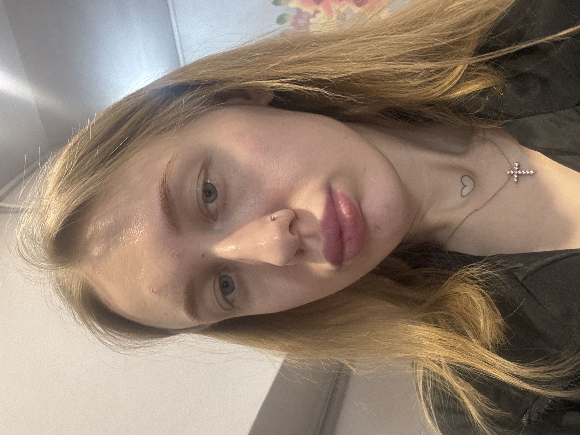 A close-up, slightly angled, portrait of a light-skinned female. Her hair is blonde and appears to be shoulder-length. She has light blue eyes and a nose piercing. A heart tattoo and a cross pendant are visible on her chest. The background is out of focus but includes a plain wall and a floral pattern. No location information for Novoaleksandrovka, Russia is provided.