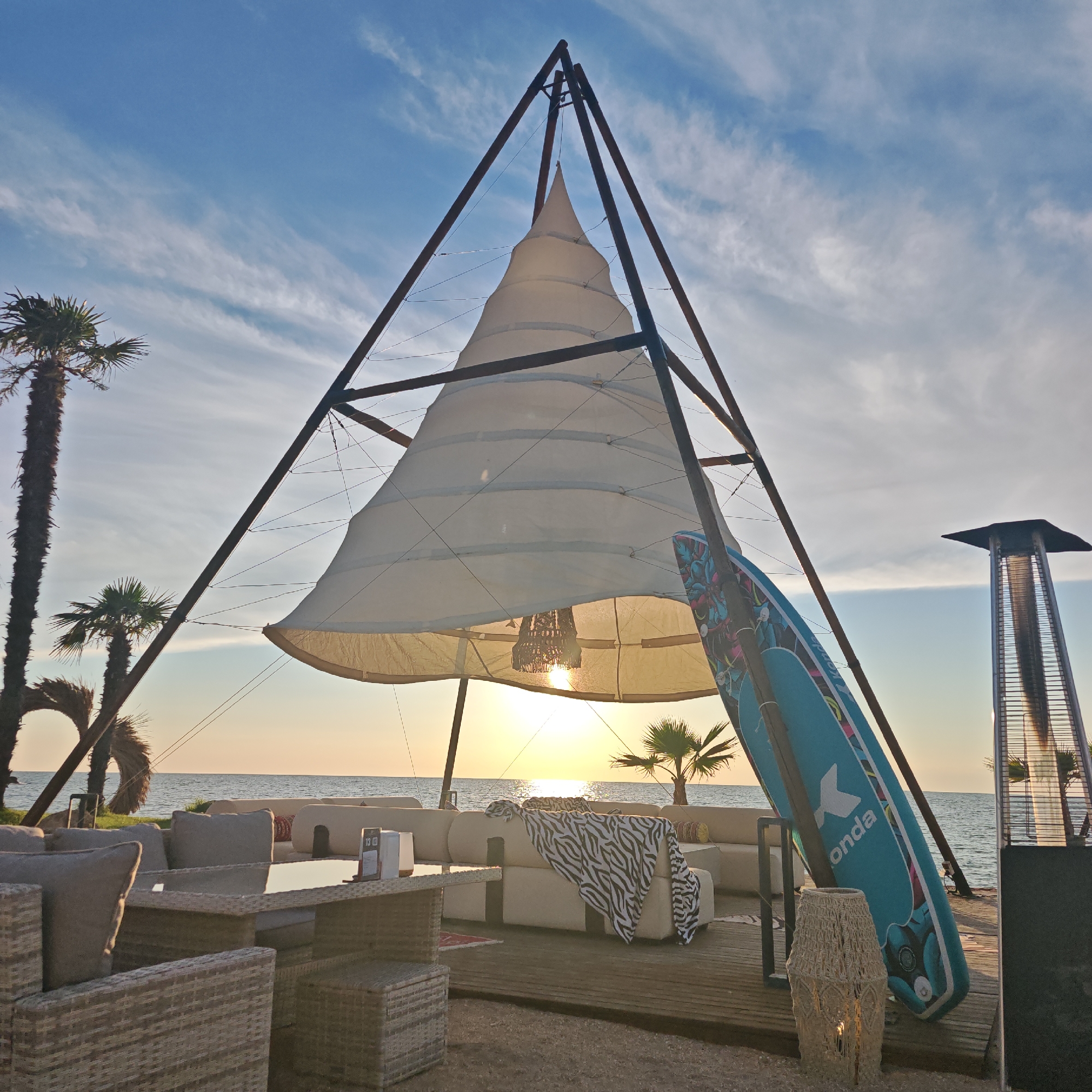 The image depicts an outdoor seating area at sunset, likely near a beach or waterfront. The scene is set in the city of Shchelkino, Ukraine. Dominating the center is a large, conical sail-like structure supported by a dark metal frame. Beneath this structure, a modern outdoor sofa with light-colored cushions is arranged, adorned with a black and white patterned throw blanket. In front of the sofa, a wicker-style coffee table with a glass top is visible, with a placemat and menus on it. To the right of the sofa, leaning against the metal frame, is a blue stand-up paddleboard with colorful graphics and the word "onda" visible. A decorative woven lantern with a light inside sits on the wooden deck in front of the paddleboard. To the far right, a tall, stainless steel patio heater with a glass column is present. In the background, the calm sea stretches to the horizon, with the setting sun casting a warm glow. Two palm trees are visible on the left, with one tall and slender, and another shorter one behind it. The sky above is a clear blue with wispy white clouds. The overall impression is one of a relaxed, seaside lounge or cafe. There are no people visible in the image.