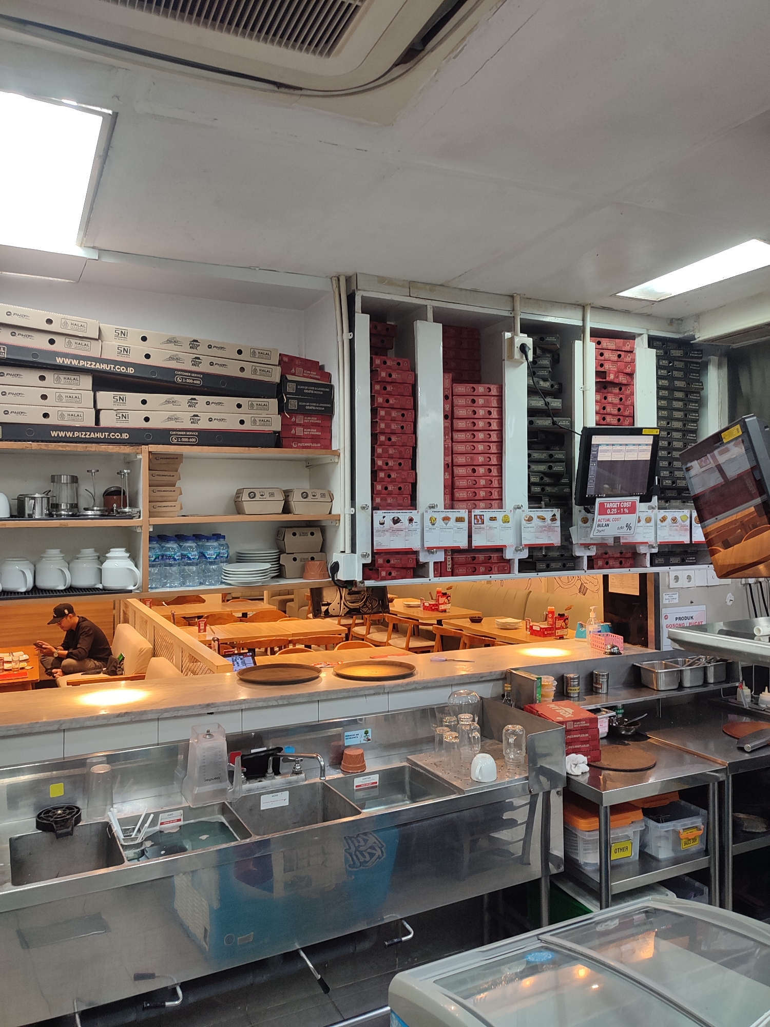 The image depicts the interior of a Pizza Hut restaurant, likely in Jember, Indonesia, given the presence of "WWW.PIZZAHUT.CO.ID" on pizza boxes. The scene is divided into a customer seating area and a service area. In the foreground, a stainless steel counter with multiple sinks is visible, equipped with faucets, soap dispensers, and cleaning supplies. Above the counter, shelves are stocked with various items, including bottled water, plates, and stacks of pizza boxes. To the left, a man wearing a black cap and black shirt is seated at a table, looking at his phone, indicating a customer is present. Behind the counter, a tall shelving unit displays numerous stacked pizza boxes, some with "HALAL" and "SNI" labels, suggesting compliance with local food and safety standards. A computer monitor is positioned on a shelf, displaying what appears to be a point-of-sale system or inventory management interface. The overall environment is clean and organized, characteristic of a fast-food establishment.