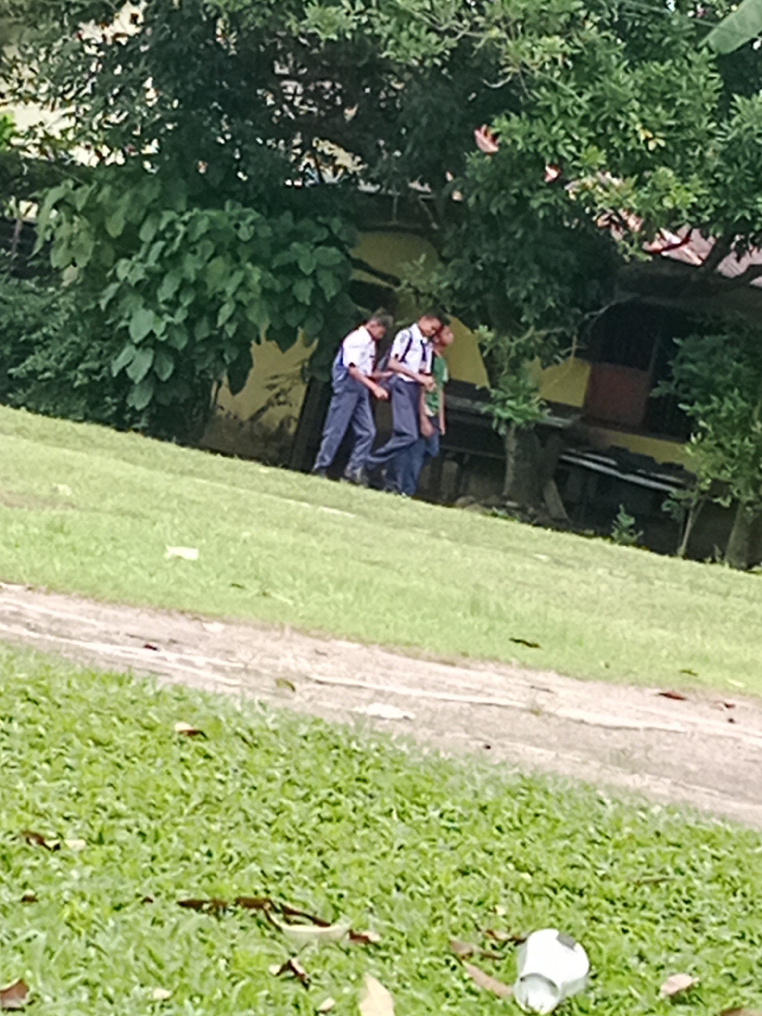 In a grassy outdoor area, two individuals wearing school uniforms are walking. One, in the foreground, is wearing a white shirt and blue pants, while the other, slightly behind and to the right, is wearing a white shirt with a dark tie and blue pants. They are walking in the same direction. The background features lush green trees and foliage, with a building structure visible behind the trees, partially obscured. The ground is covered in grass, with a dirt path running diagonally across the lower portion of the image. A white object, possibly a discarded cup, is visible on the grass in the lower right corner. The scene appears to be in a park or school grounds in the City of Binjai, Indonesia.
