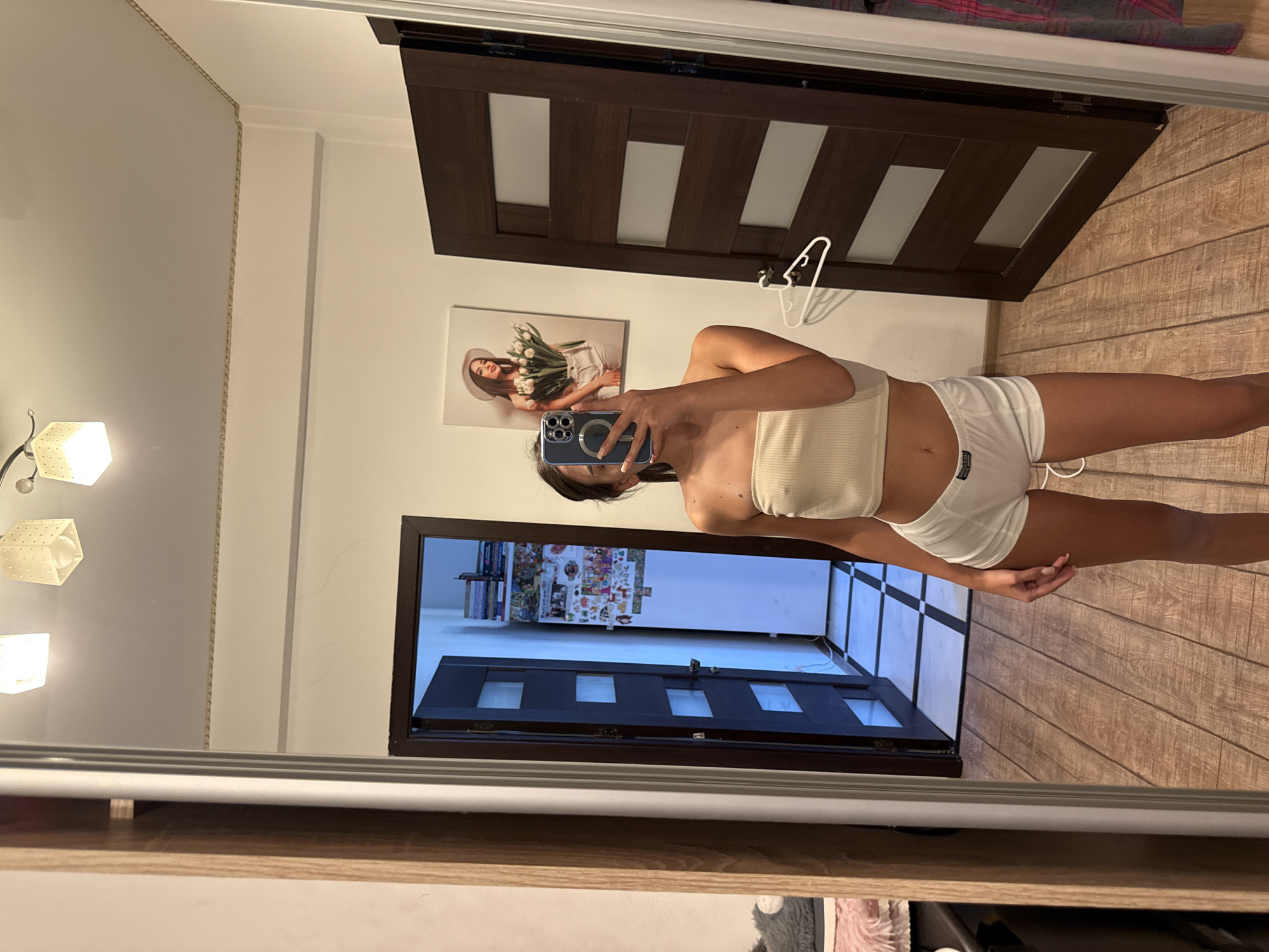 A woman is standing in front of a mirror, taking a selfie. She is wearing a light yellow tube top and white shorts. Behind her, to the left, is a door with glass panels, and to the right, another dark wooden door. On the wall, there is a framed picture of a woman holding tulips. The room has a wooden floor and white walls. The overall scene suggests an indoor environment, possibly a bedroom or dressing area, located within an apartment in Chișinău, Moldova.