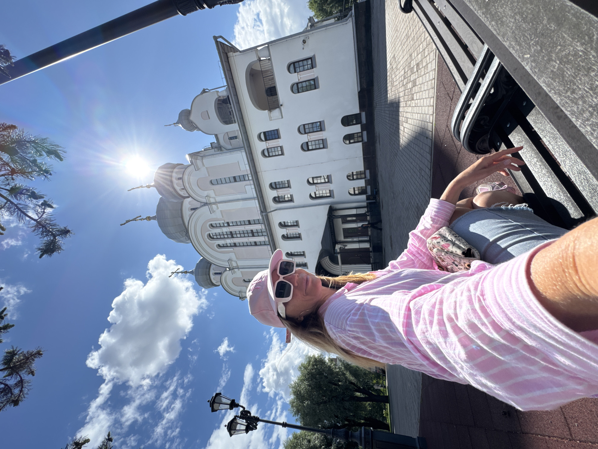 A woman with blonde hair is sitting on a park bench in front of a white church. She is wearing a pink and white striped shirt, a pink baseball cap, white sunglasses, and blue jeans. The church has a white facade with a pink trim and several domes. The sky is blue with white clouds, and the sun is shining brightly. There is a lamppost to the left of the woman. The image appears to be taken in Saint Petersburg, Russia.