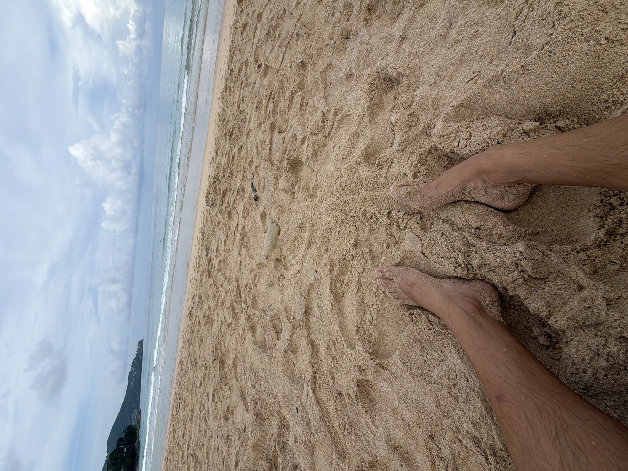 The image is a POV shot of a person's legs and feet on a sandy beach in Choeng Thale, Thailand. The sand is light brown and appears damp in some areas. The person's legs are hairy and have some water droplets visible on them. Their feet are partially buried in the sand. In the background, the ocean meets the sky under a cloudy day. There are gentle waves rolling onto the shore. The shoreline extends to the left, where a lush green hillside with some buildings is visible. Two objects, possibly a bottle and a piece of dark debris, are present in the sand further up the beach. The overall scene depicts a relaxed moment on a tropical beach.