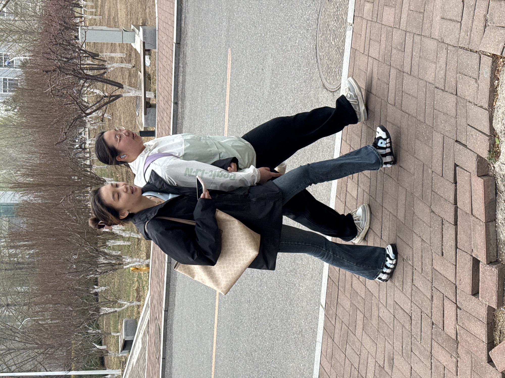 This media file depicts two young Asian women walking side-by-side on a brick sidewalk next to an asphalt road, suggesting an urban or campus setting in Zhongtun, China. The scene appears to be in late winter or early spring, indicated by the bare trees that line a grassy area adjacent to the road.
The woman on the left is wearing a black jacket, blue jeans, and black and white striped sneakers. She carries a light-colored, patterned shoulder bag and is holding a smartphone. Her hair is styled in two buns, and she has a pleasant, relaxed expression.
The woman on the right is dressed in a light green and white jacket with the text "Donk-N" visible on its sleeve, black pants, and light-colored sneakers. She wears glasses and earbuds, and also appears to be holding a smartphone. She has a subtle smile on her face.
The lighting is bright but diffused, suggesting an overcast daytime sky. In the background, beyond the leafless trees (some marked with white bands on their trunks), faint outlines of buildings can be seen. A manhole cover is visible on the asphalt road. Both women seem to be enjoying a casual stroll outdoors.