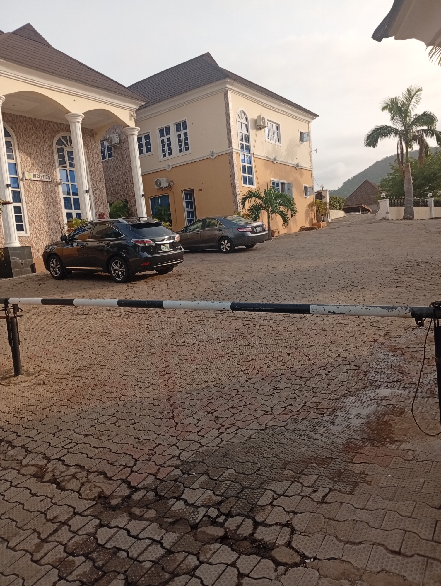 The image depicts the exterior of a building with a "RECEPTION" sign, likely a hotel or guesthouse in Lokoja, Nigeria. A black SUV and a grey sedan are parked in the paved driveway. A black and white security barrier is in the foreground, partially obscuring the view. The paving stones are reddish-brown and patterned. In the background, there is another building and a palm tree. A hill is visible in the distance. The sky is overcast. There are no people visible in the image.