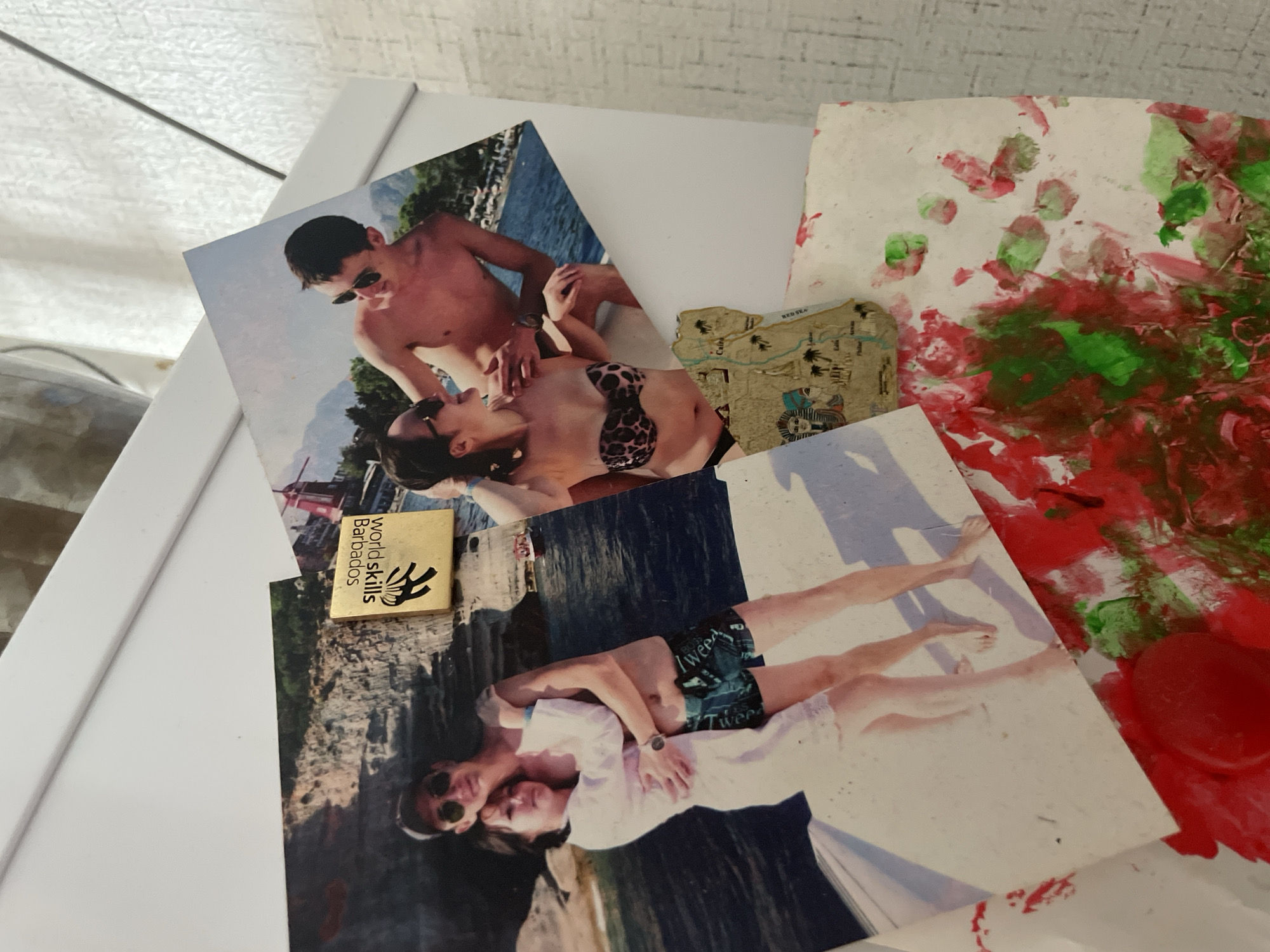 The image shows a close-up view of a white refrigerator door, adorned with several photographs, a small gold-colored pin, and a piece of paper with painted smudges. One photograph features a couple embracing on what appears to be a boat, with a scenic background of cliffs and water. The man is wearing sunglasses and patterned swim trunks, while the woman is in a white cover-up. Another photograph displays a couple in swimwear, with the woman in a leopard-print bikini top and the man wearing sunglasses. In the background of this photo, there are mountains and buildings. A small, square pin with an emblem and text reading "World Skills Barbados" is attached to one of the photographs. Below the photographs, a piece of paper with smudges of red and green paint is visible, possibly from a child's artwork. A small map fragment depicting Egypt is also present. The overall environment suggests a personal display area, likely in a home. There is no visible indication of the location being within the city of Laishevo, Russia.
