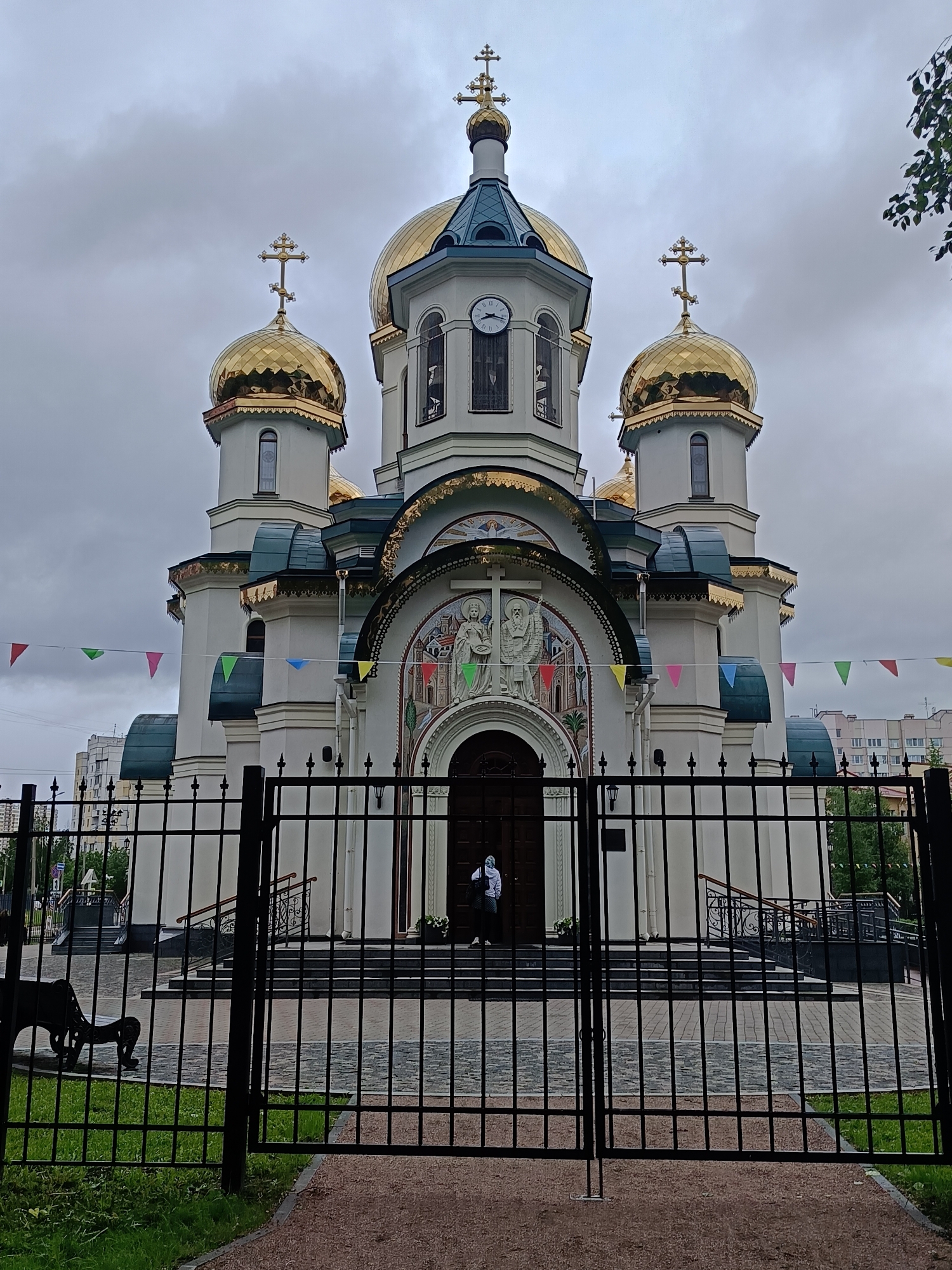 A medium shot captures the Cathedral of the Icon of the Mother of God "Joy of All Who Sorrow" in Saint Petersburg, Russia. The ornate white and gold cathedral, enclosed by a black iron fence, features multiple golden domes topped with Orthodox crosses. A person stands in the doorway, partially obscured. The scene is set on a cloudy day, with colorful flags strung between the cathedral's structures. A wrought-iron bench is visible on the lawn behind the fence, and buildings are seen in the background. The gate is shut and in the foreground of the picture.