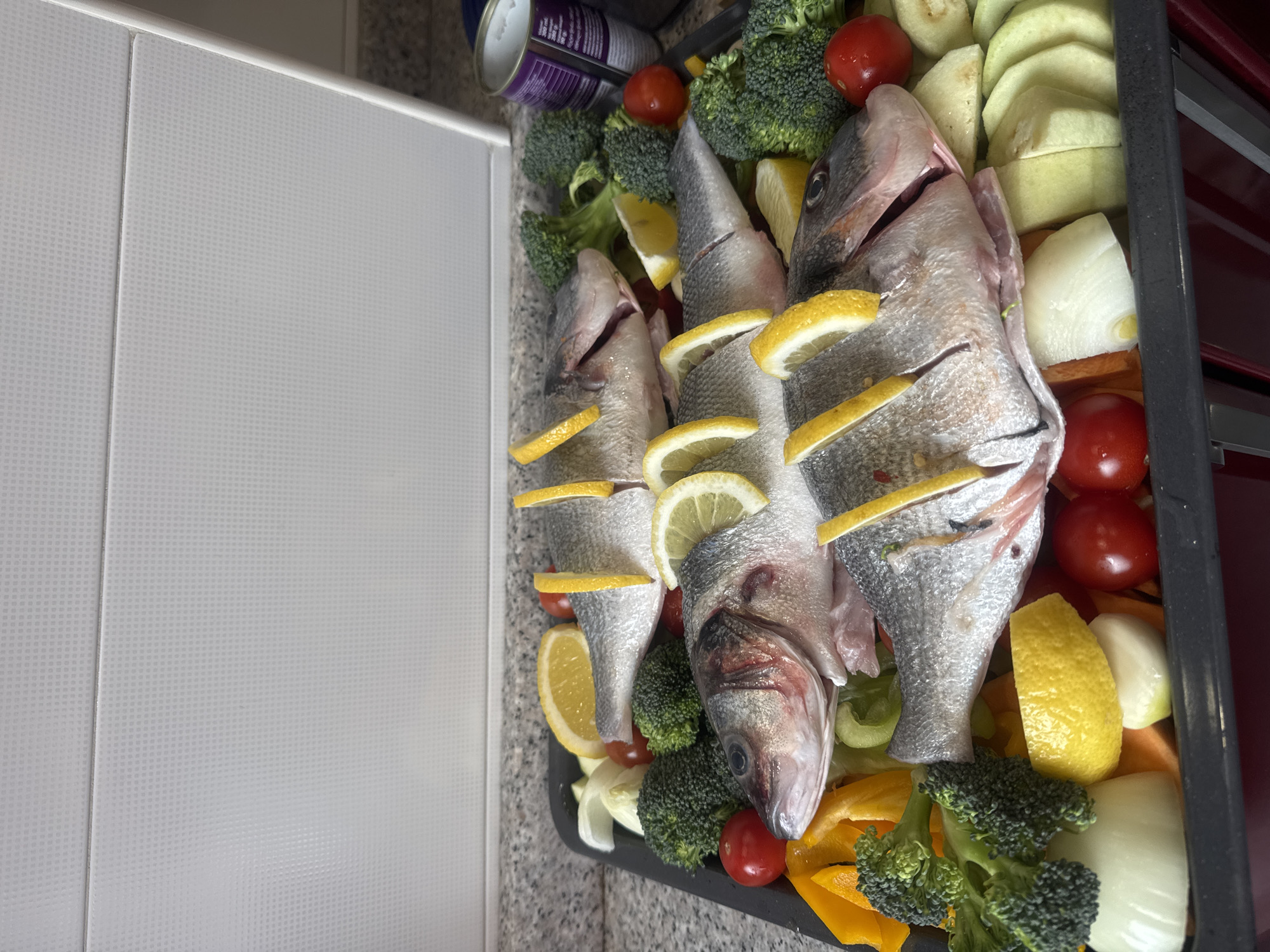 The image shows a black baking tray filled with three whole fish, interspersed with lemon slices, broccoli florets, cherry tomatoes, sliced squash or zucchini, and chopped onions and bell peppers. A can is also visible in the upper left. The tray appears to be on a kitchen countertop. The location is not discernible from the image.